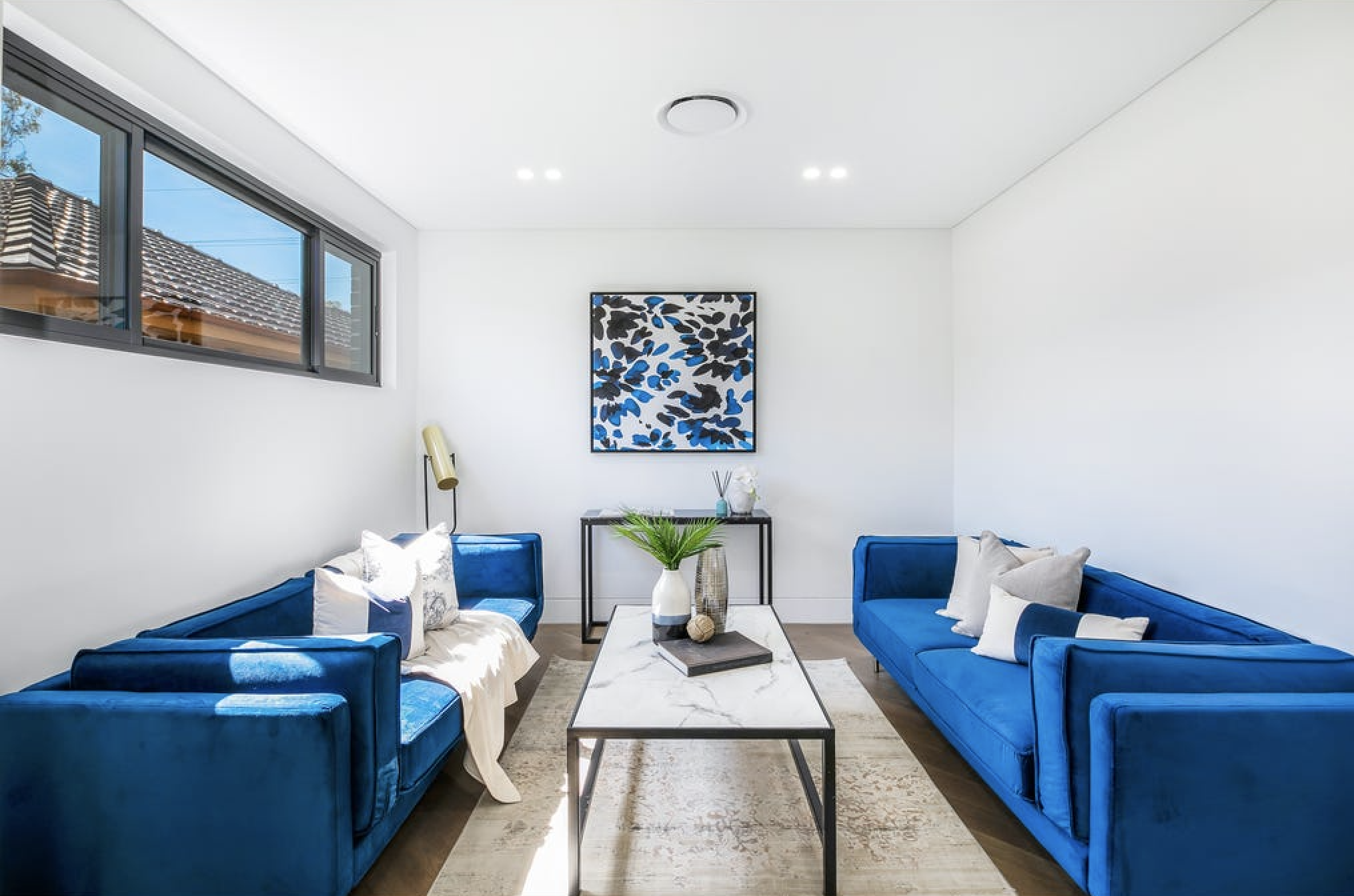 Contemporary living room with two bright blue velvet sofas, a marble coffee table, a minimalist console table with decorative items, a large window, and a framed abstract art piece on the white wall.