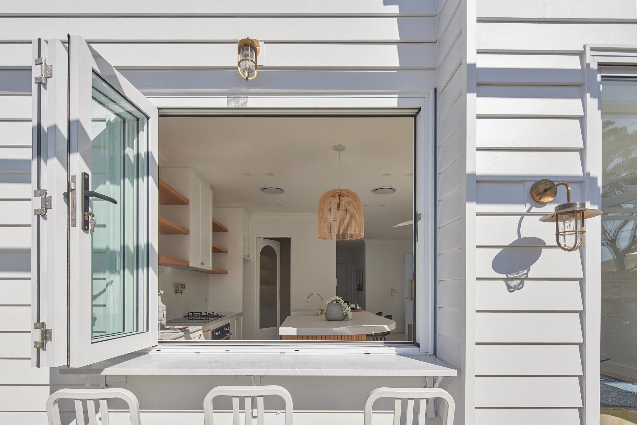 Open window of a white house revealing modern kitchen with countertop, open shelves, and hanging woven light fixture.