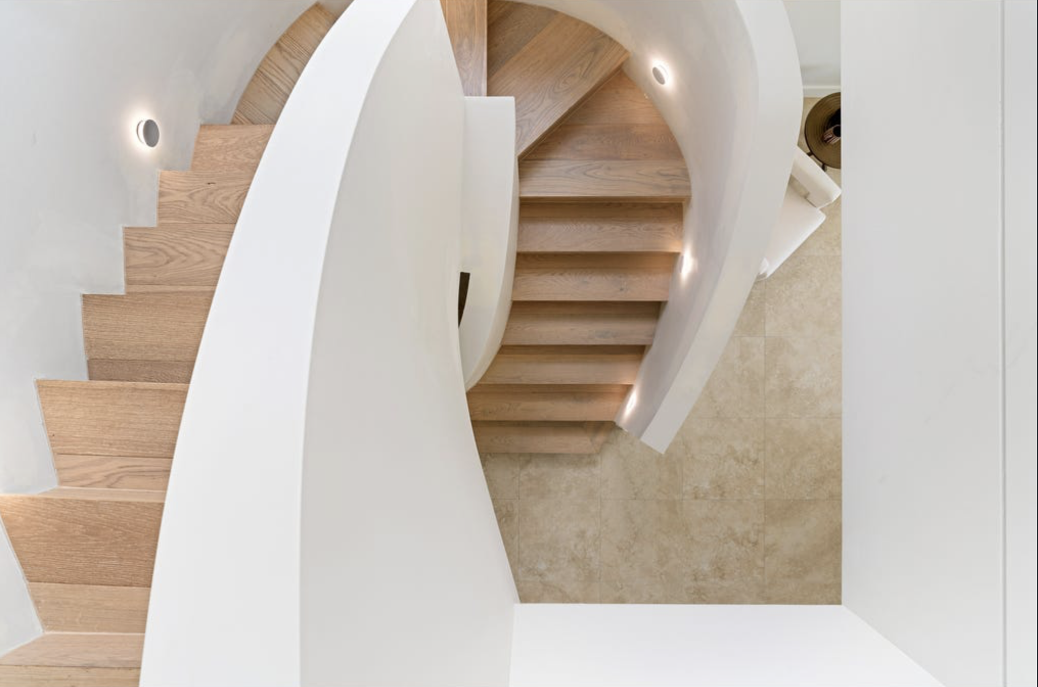 Top-down view of a modern staircase with wooden steps and white walls, featuring concealed lighting along the curved wall beside the stairs.
