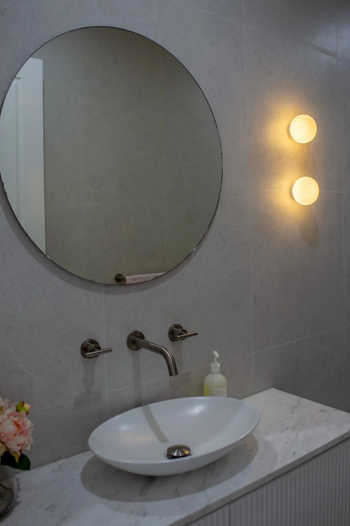 A modern bathroom mirror above a white sink with a soap dispenser on a marble countertop, featuring wall-mounted lighting and gray tiled walls.