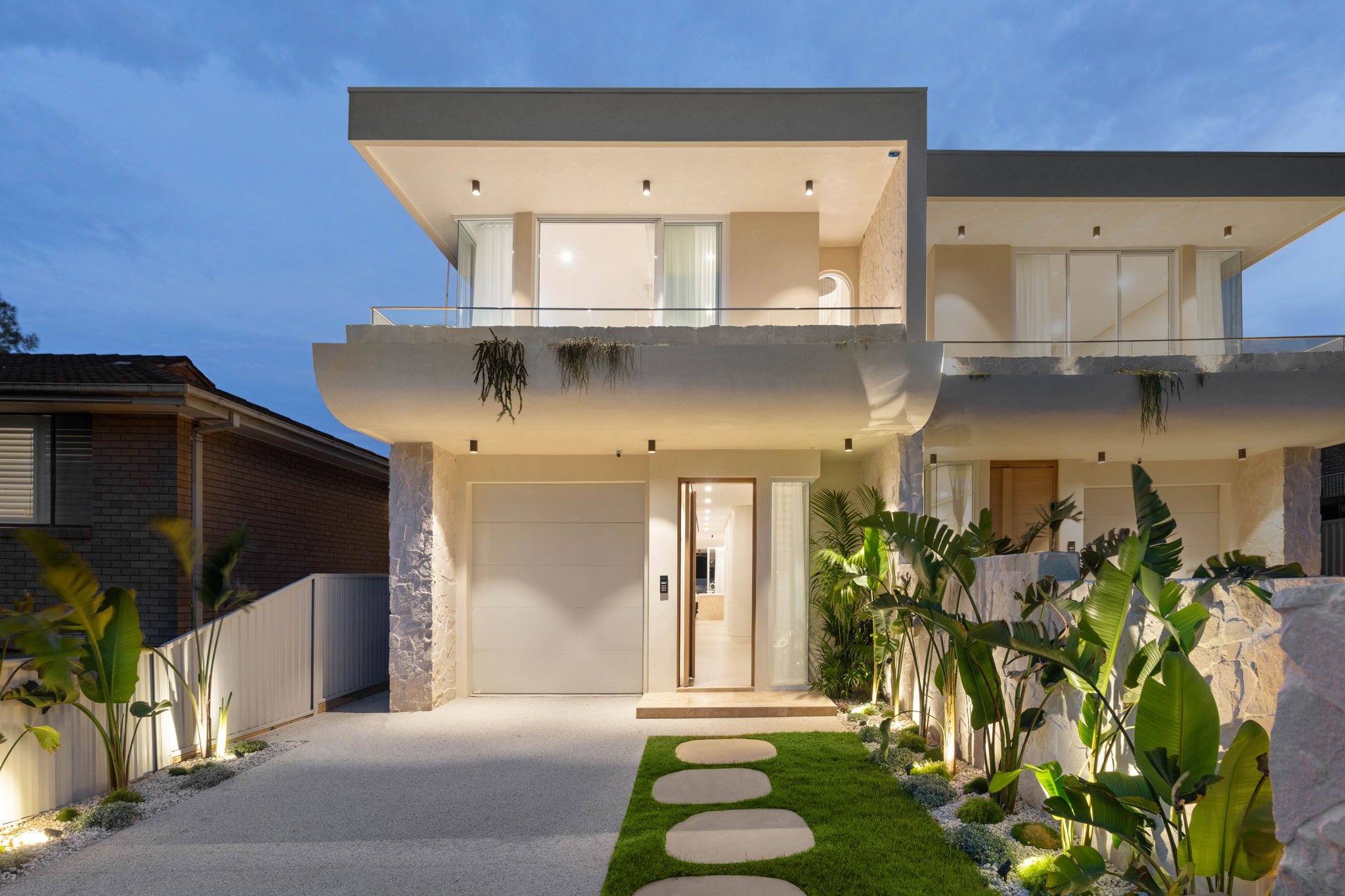 Modern two-story house with white exterior, illuminated exterior lights, and landscaping featuring tall green plants and decorative stones, during dusk.