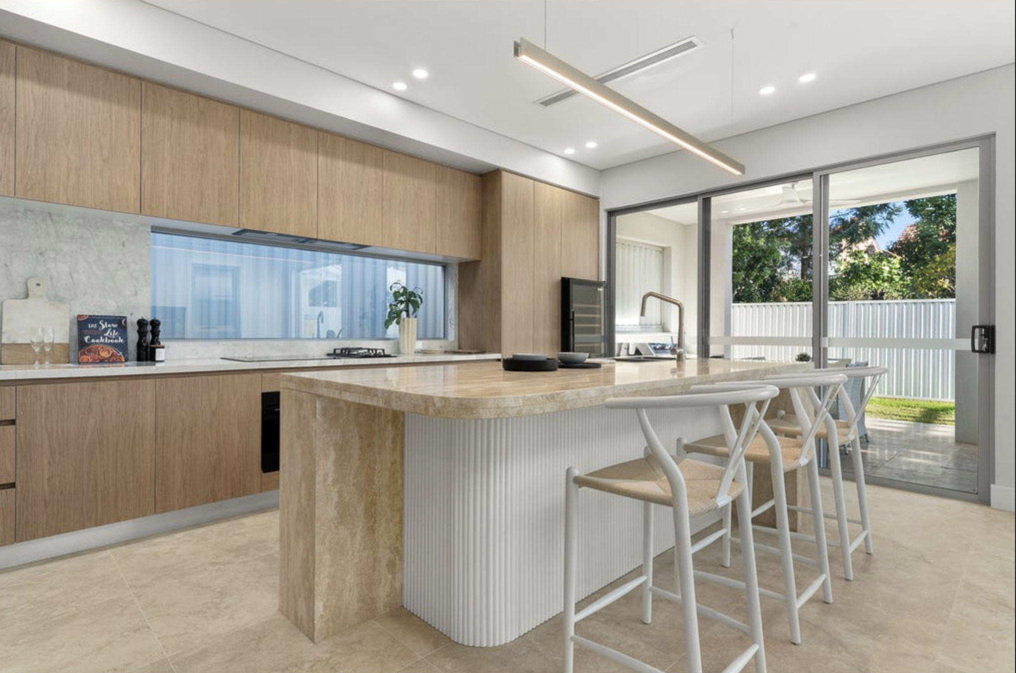 Modern kitchen with wooden cabinets, marble island, beige bar stools, and large sliding glass door leading to an outdoor yard.