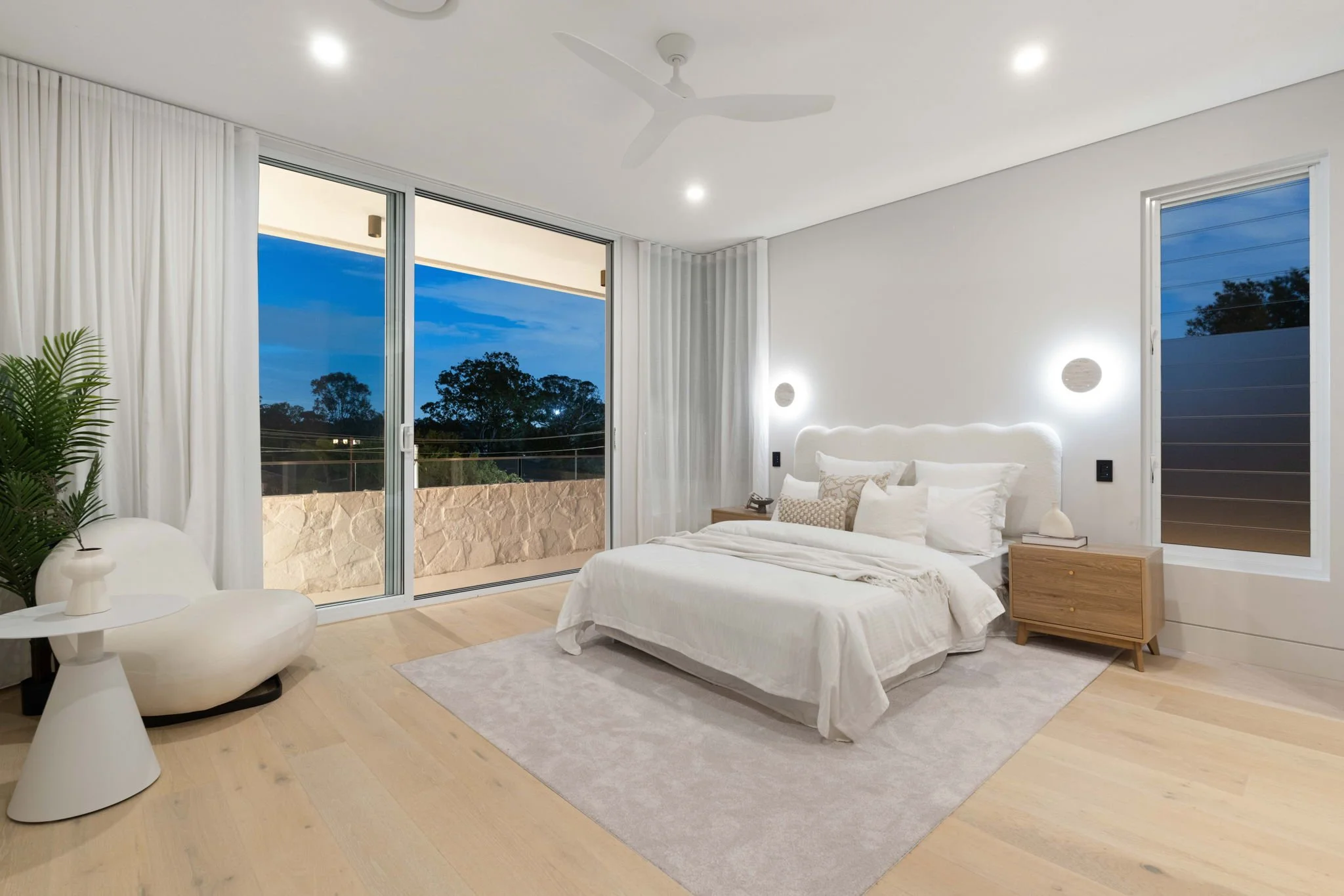 Modern bedroom with white walls, large sliding glass door showing outdoor balcony, and a window. A bed with white bedding and pillows, nightstands on each side with lamps, and a ceiling fan. Light wood flooring and a light-colored area rug.