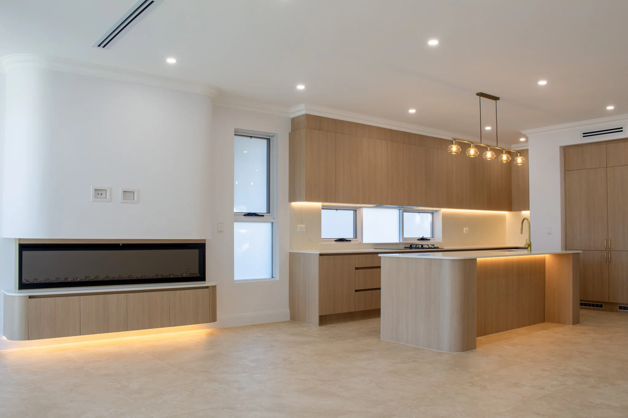 Modern kitchen with light wood cabinets, a kitchen island, a gas stove, a brass faucet, and a built-in electric fireplace in the living area. Recessed lighting and a hanging chandelier provide illumination.