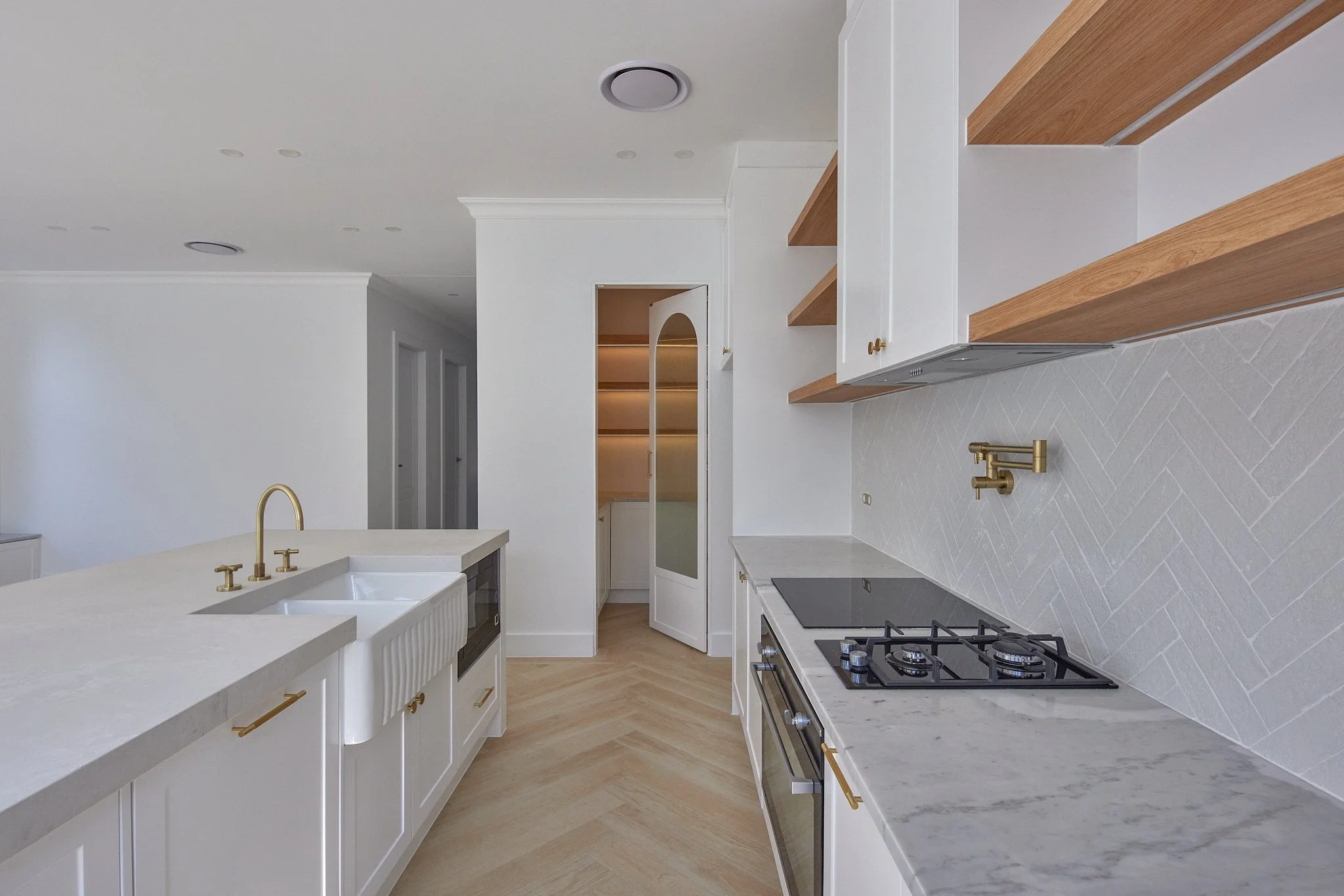 Modern kitchen with white cabinets, marble countertops, a gold faucet, a black gas stove, open wooden shelves, and a white backsplash with a chevron pattern.