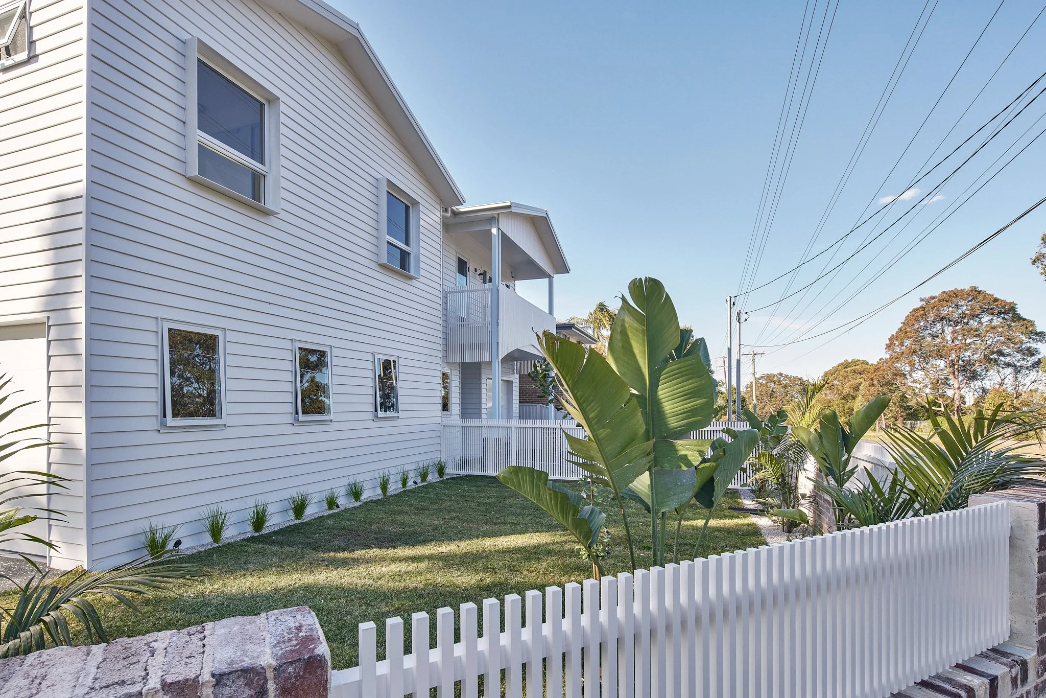 A white two-story house with multiple windows, a small lawn with tropical plants, a white picket fence, and power lines in the background under a clear sky.