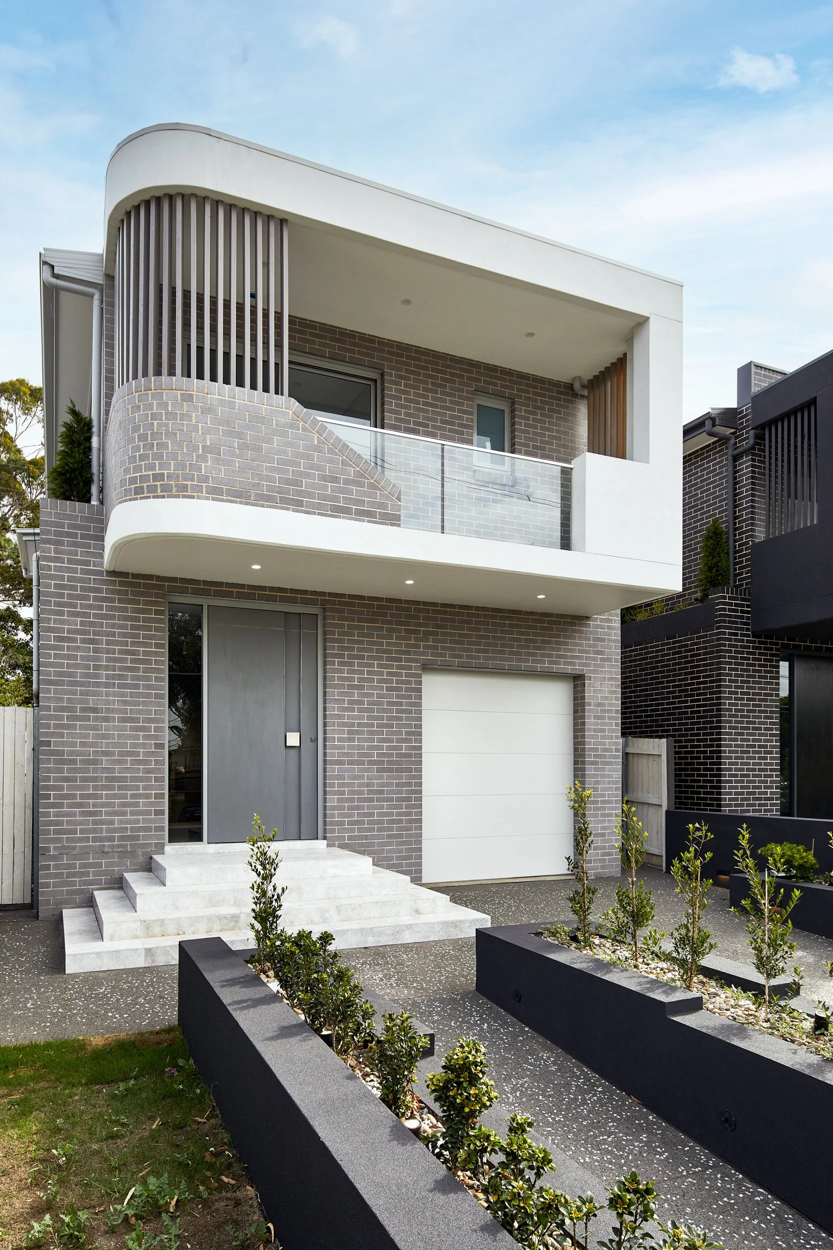 Modern two-story house with a brick facade, white balcony, gray front door, and a small front yard with planting beds and a concrete pathway.