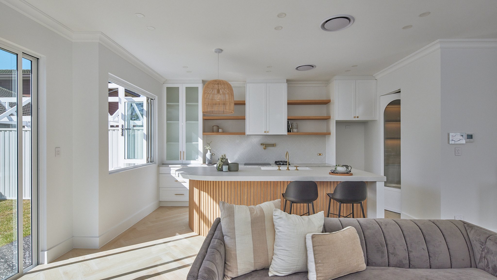 Modern open-concept kitchen with white cabinetry, wooden open shelves, and a central island with a wooden panel. Sunlight streams through large windows, illuminating the space and adjacent living area with a gray sofa and beige pillows.