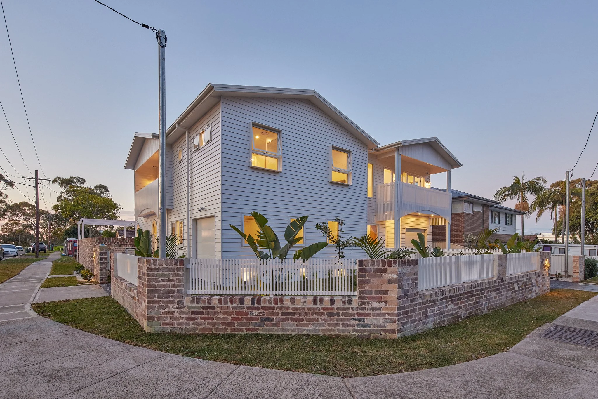 A modern two-story house with white siding, surrounded by a brick and white picket fence, with palm trees and a sidewalk in the neighborhood at dusk.
