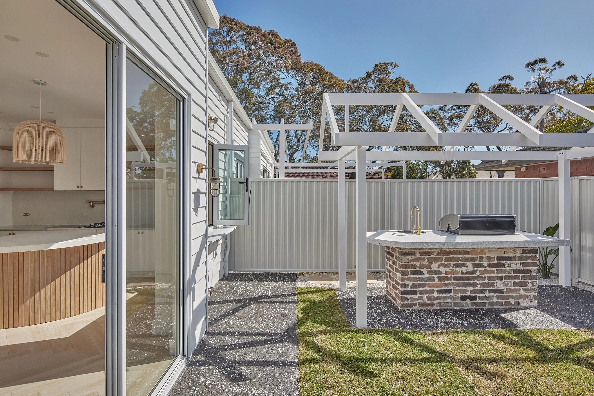 Outdoor backyard area with a pergola frame, a brick outdoor kitchen counter with a grill, sliding glass door, and open window showing inside kitchen with pendant light and cabinetry.