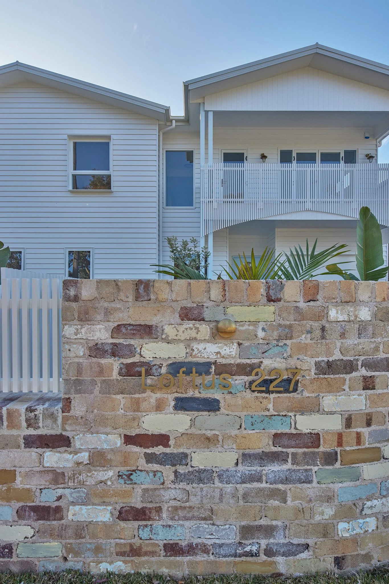Brick wall with a sign that reads "Loftus 227" in front of a white residential building with multiple windows, a balcony, and tropical plants.