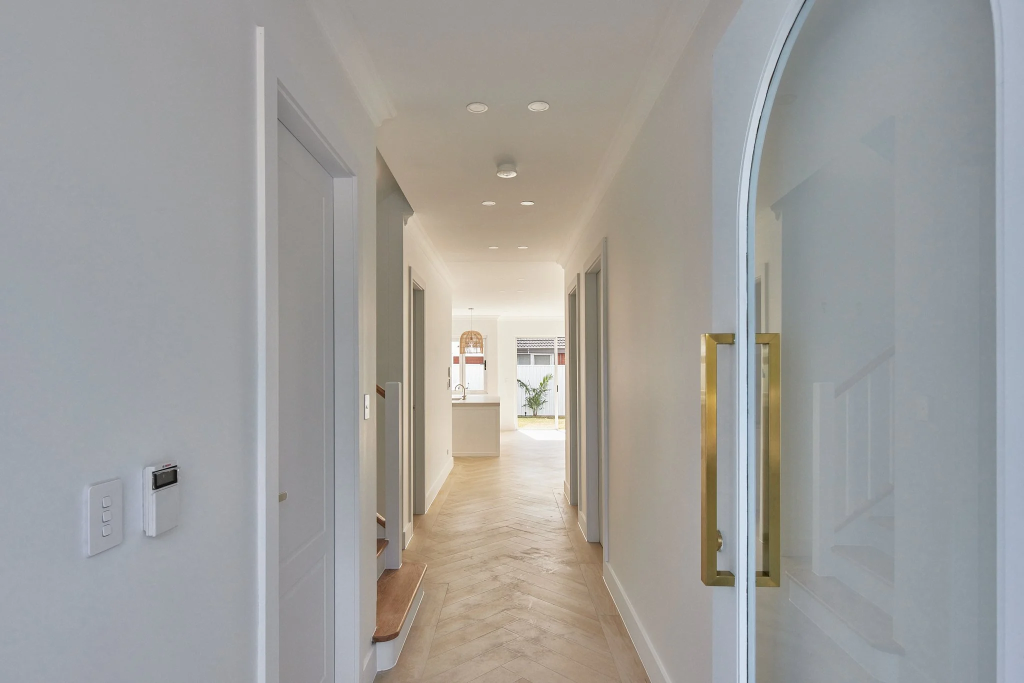 Bright hallway in a modern home with white walls, wooden flooring, and doors on both sides leading to different rooms. There are recessed ceiling lights, and at the end, there is a glimpse of a kitchen and a door leading outside with a small yard.