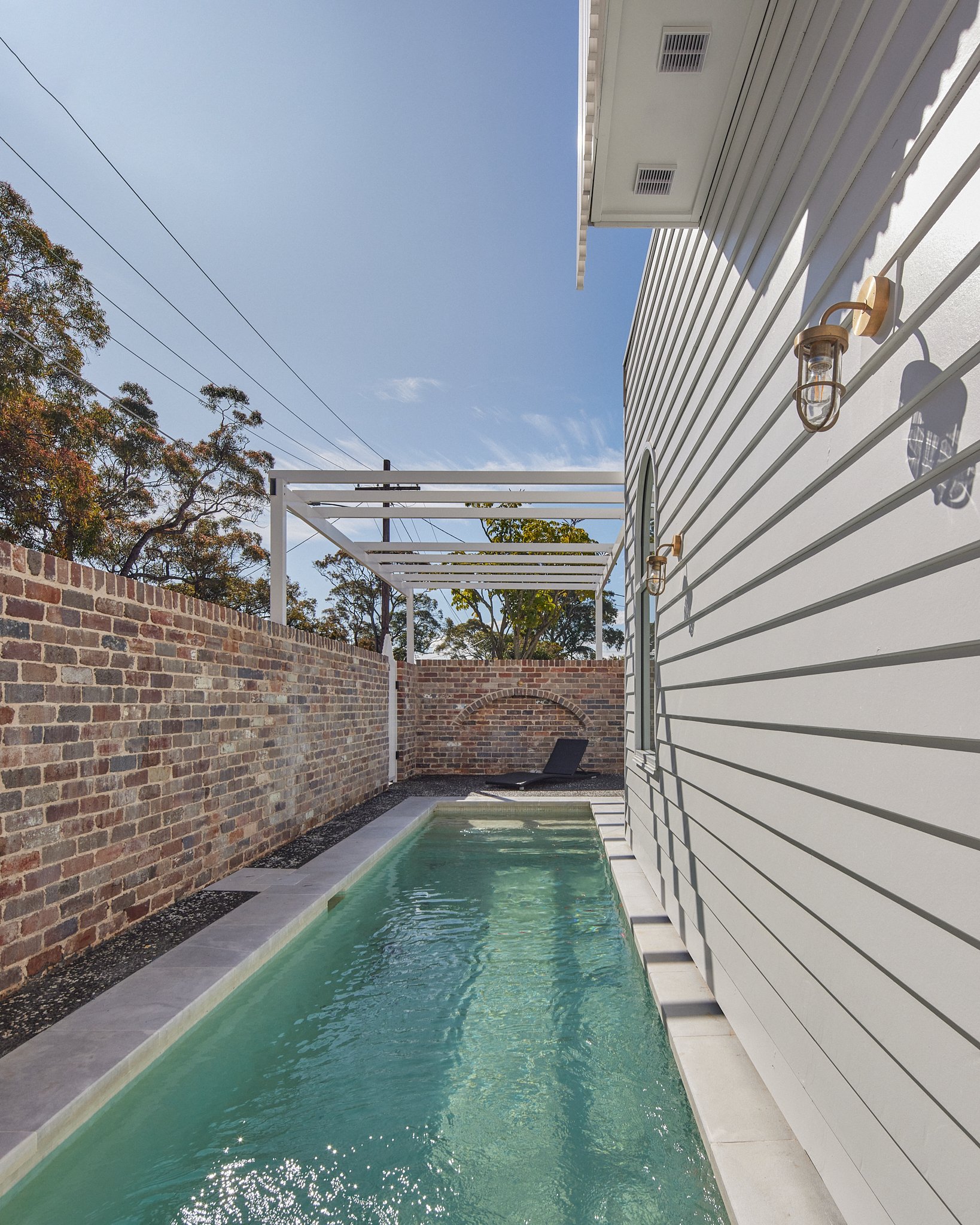 A narrow swimming pool alongside a white house with siding and outdoor lighting fixtures, enclosed by a brick wall, with a lounge chair at the far end and green trees in the background under a partly cloudy sky.