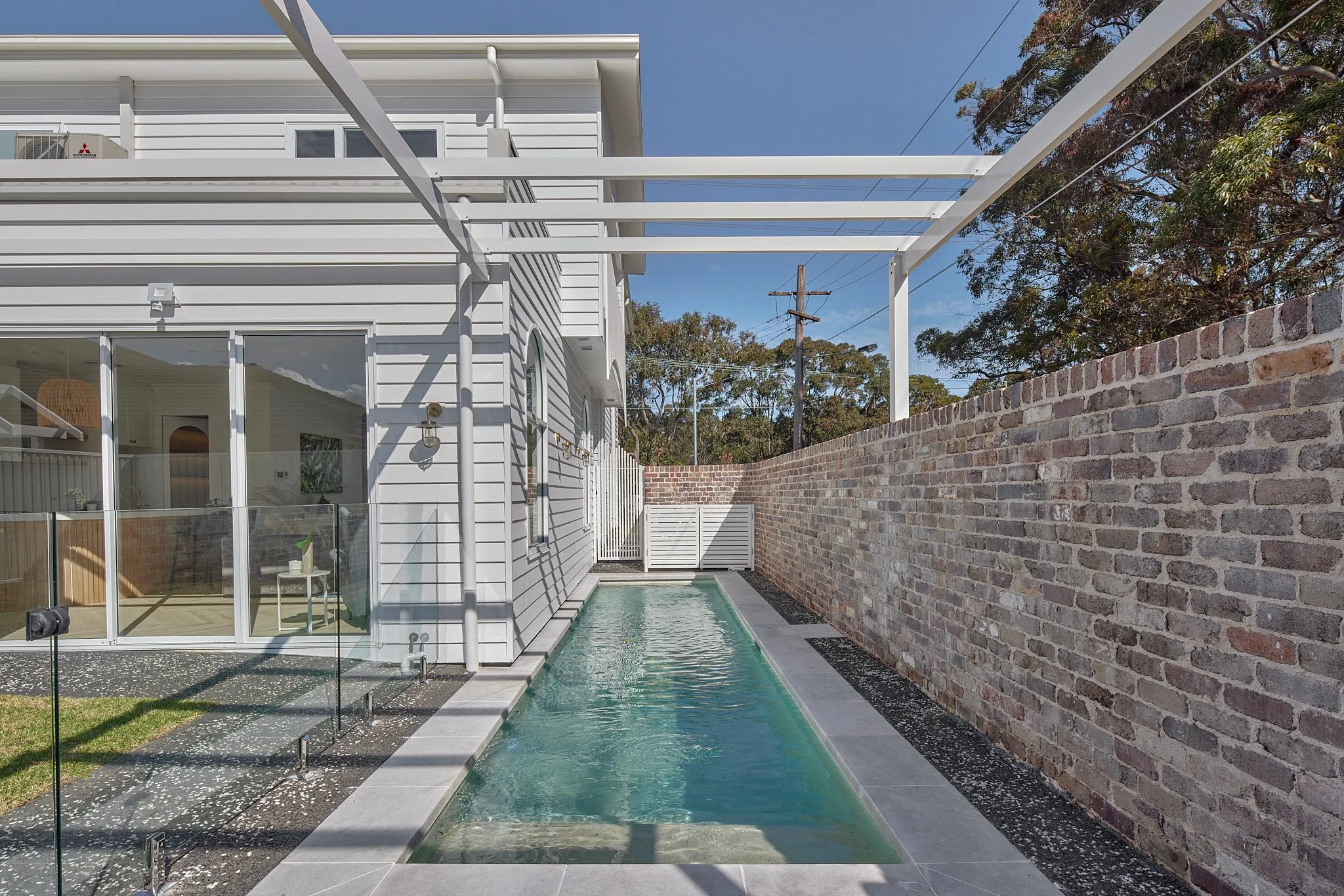 A narrow backyard with a modern white house, small rectangular pool, brick wall, and a white trellis above.