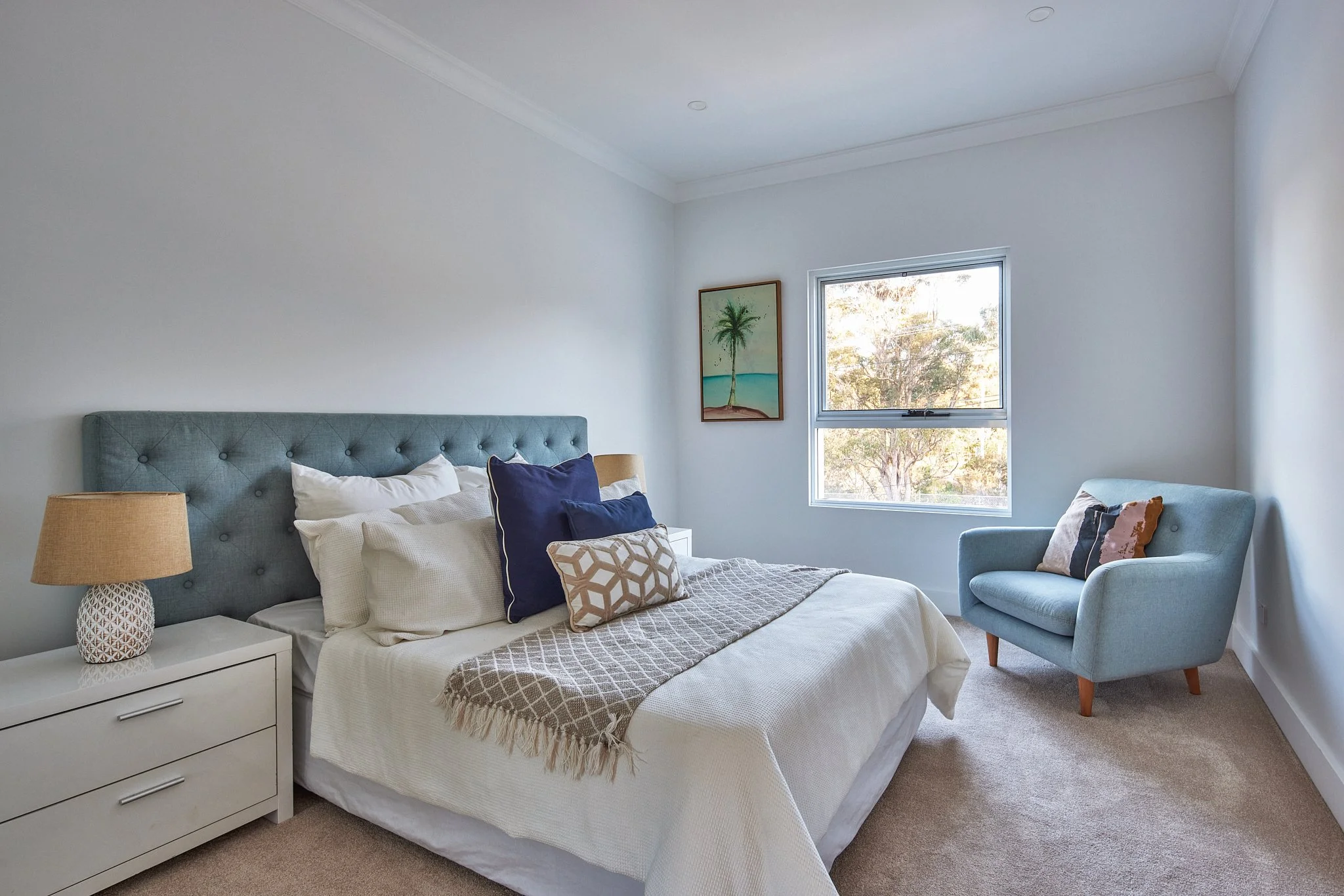 A bedroom with a white bed, blue upholstered headboard, and multiple pillows, a white nightstand with a beige lamp, a window with trees outside, a framed palm tree on the wall, and a light blue armchair with pillows.