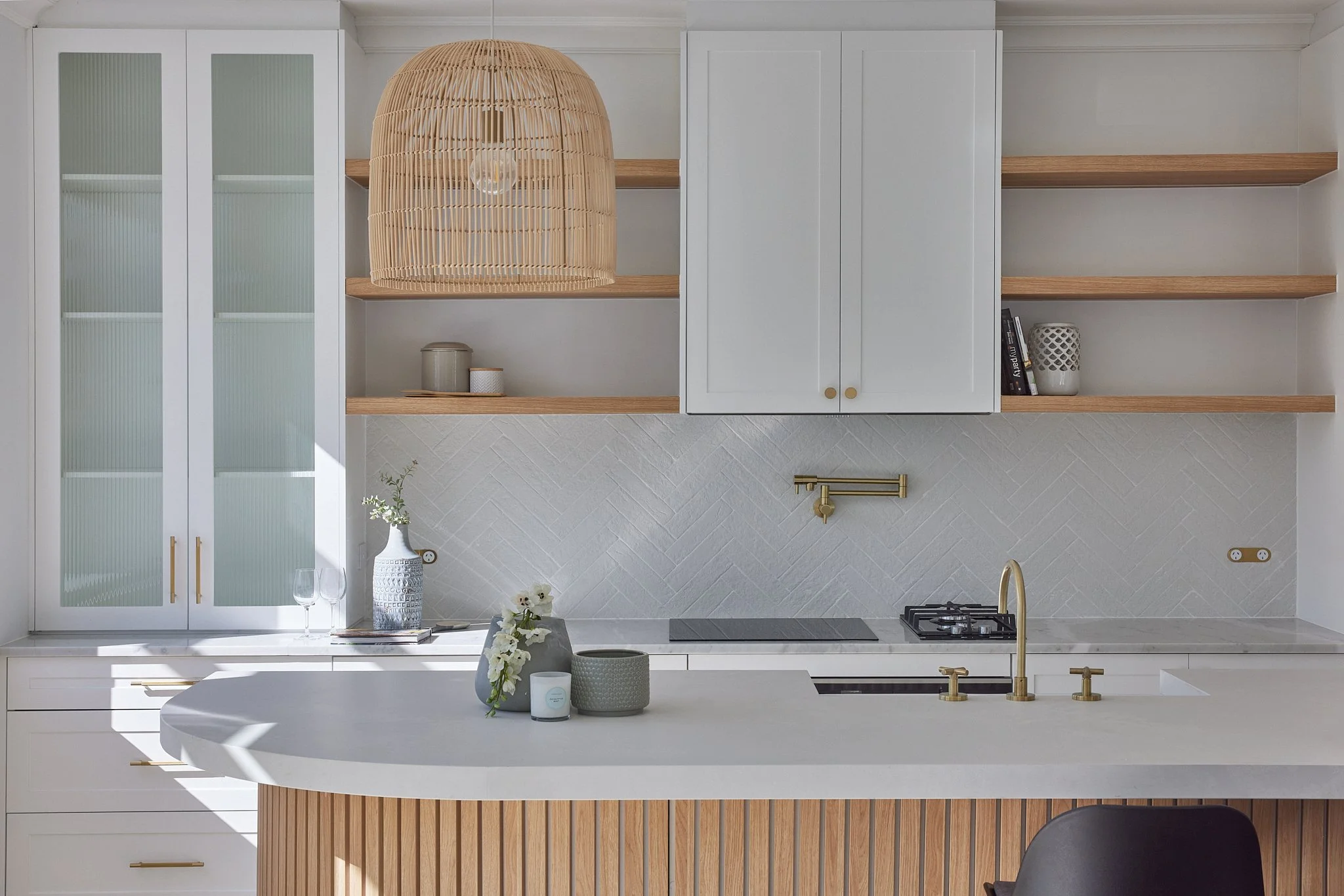 Modern kitchen with white cabinets, open wooden shelves, a large hanging rattan pendant light, and a white countertop island with gold fixtures and decorative vases.