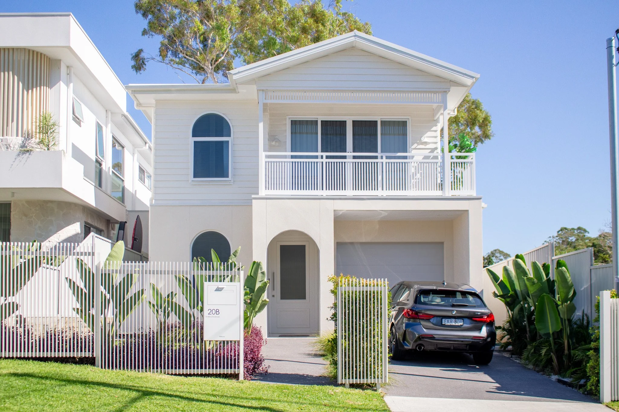 Modern two-story white house with a small front yard, a driveway with a black SUV, and a white fence. The house has a balcony with white railings and large windows.