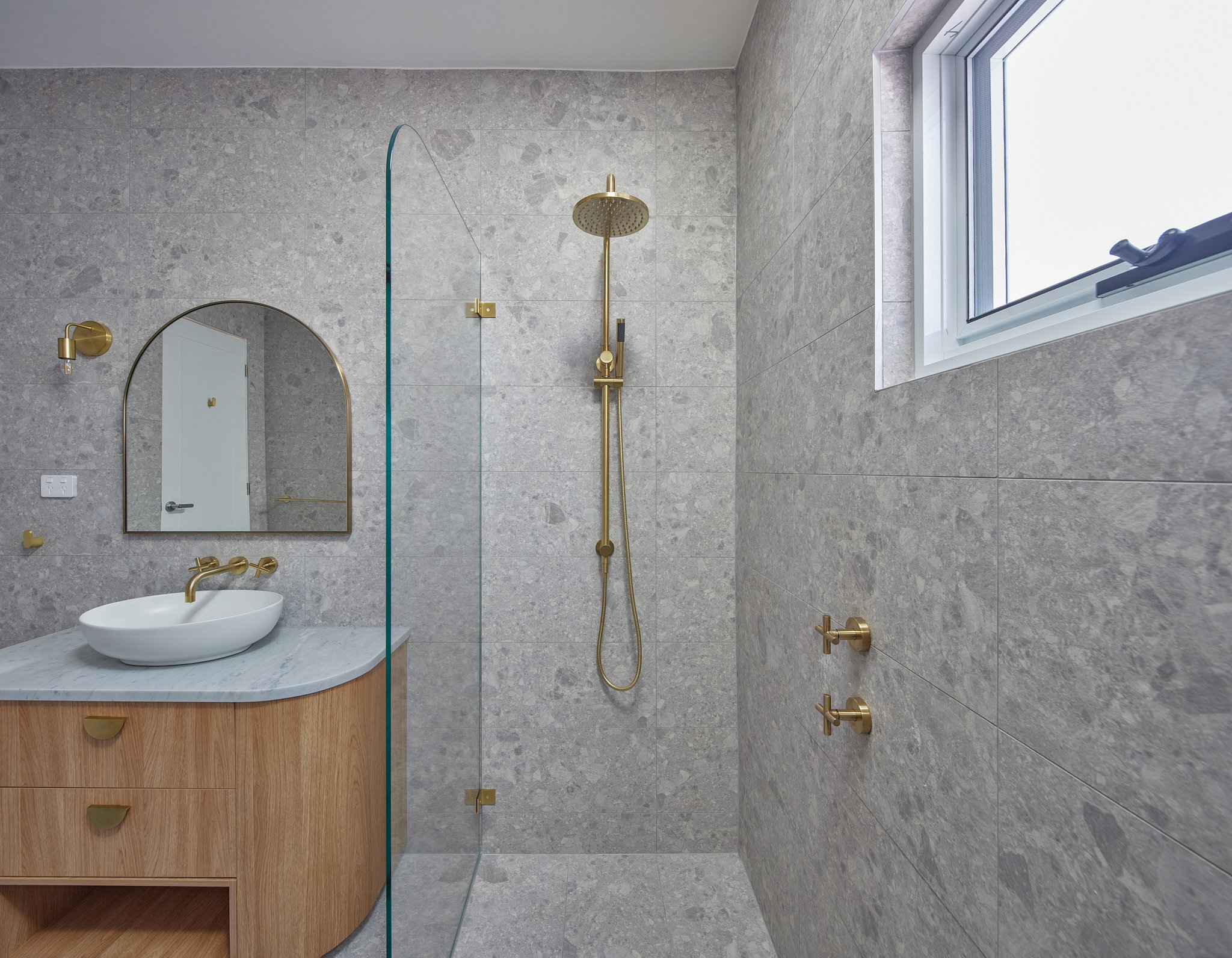 Modern bathroom with gray stone tiles, gold fixtures, a corner vanity with a vessel sink, a arched mirror, a wall-mounted light, a shower with a gold rainfall showerhead and a glass partition, and a window.