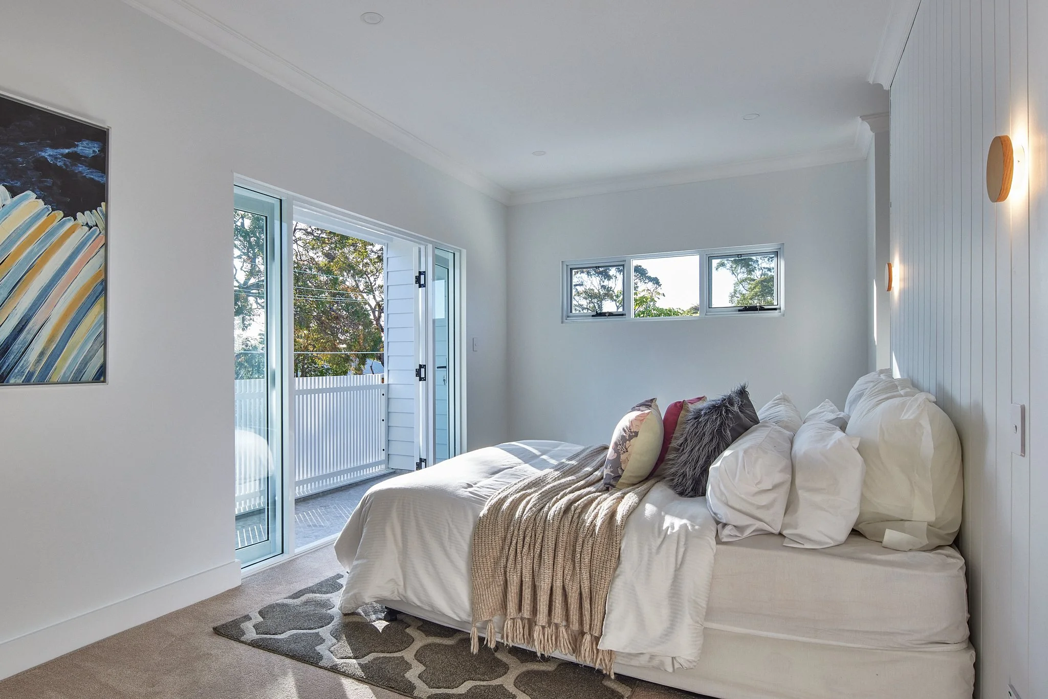 Bright bedroom with a white bed, numerous pillows, and a beige throw blanket. A sliding glass door leads to a balcony or patio, and a small window is on the wall. A colorful abstract painting is on the wall, and a patterned rug sits beneath the bed.