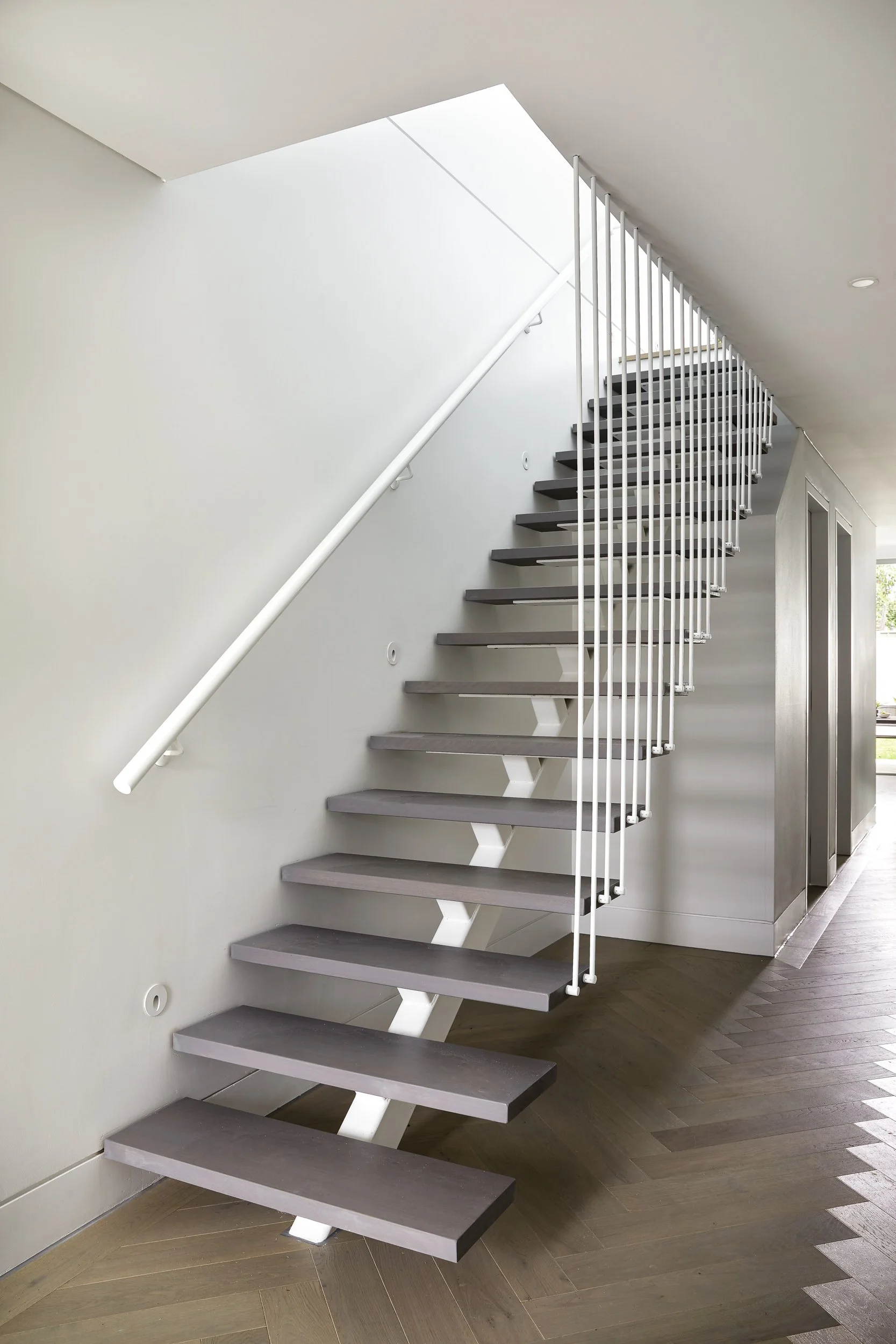 Interior view of a modern staircase with open risers, gray treads, a white handrail, and white vertical balusters, next to an elevator door, with wood flooring and white walls.
