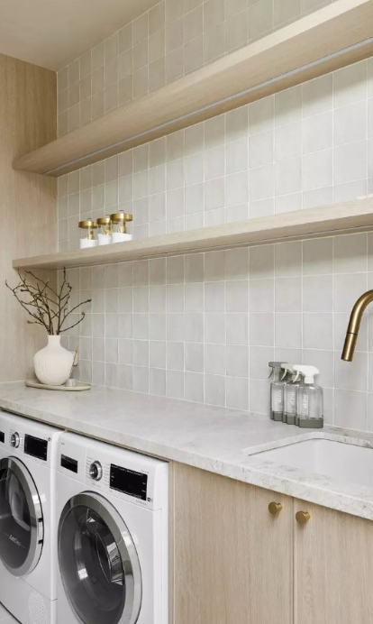 Modern laundry room with white washer and dryer, beige cabinets, and open beige shelves with decorative items, a white vase with branches, a sink, and clear soap dispensers.