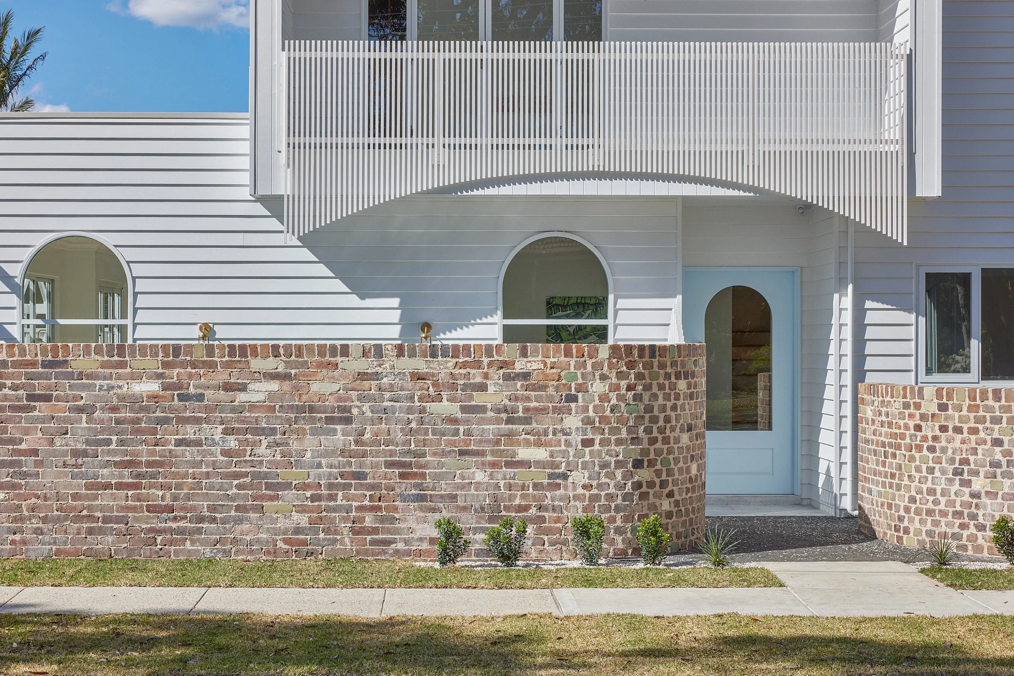 Front view of a modern white house with a brick wall and small landscaping in front, blue door, and a balcony with slatted white railing.