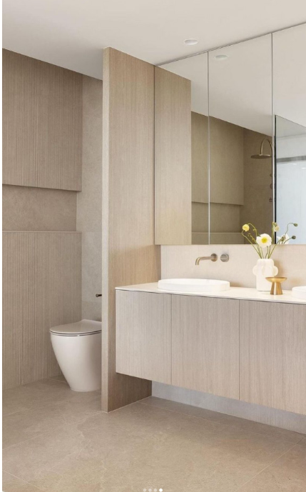 Modern bathroom with beige walls, a toilet, and a vanity with a sink and a large mirror, decorated with a vase of flowers.