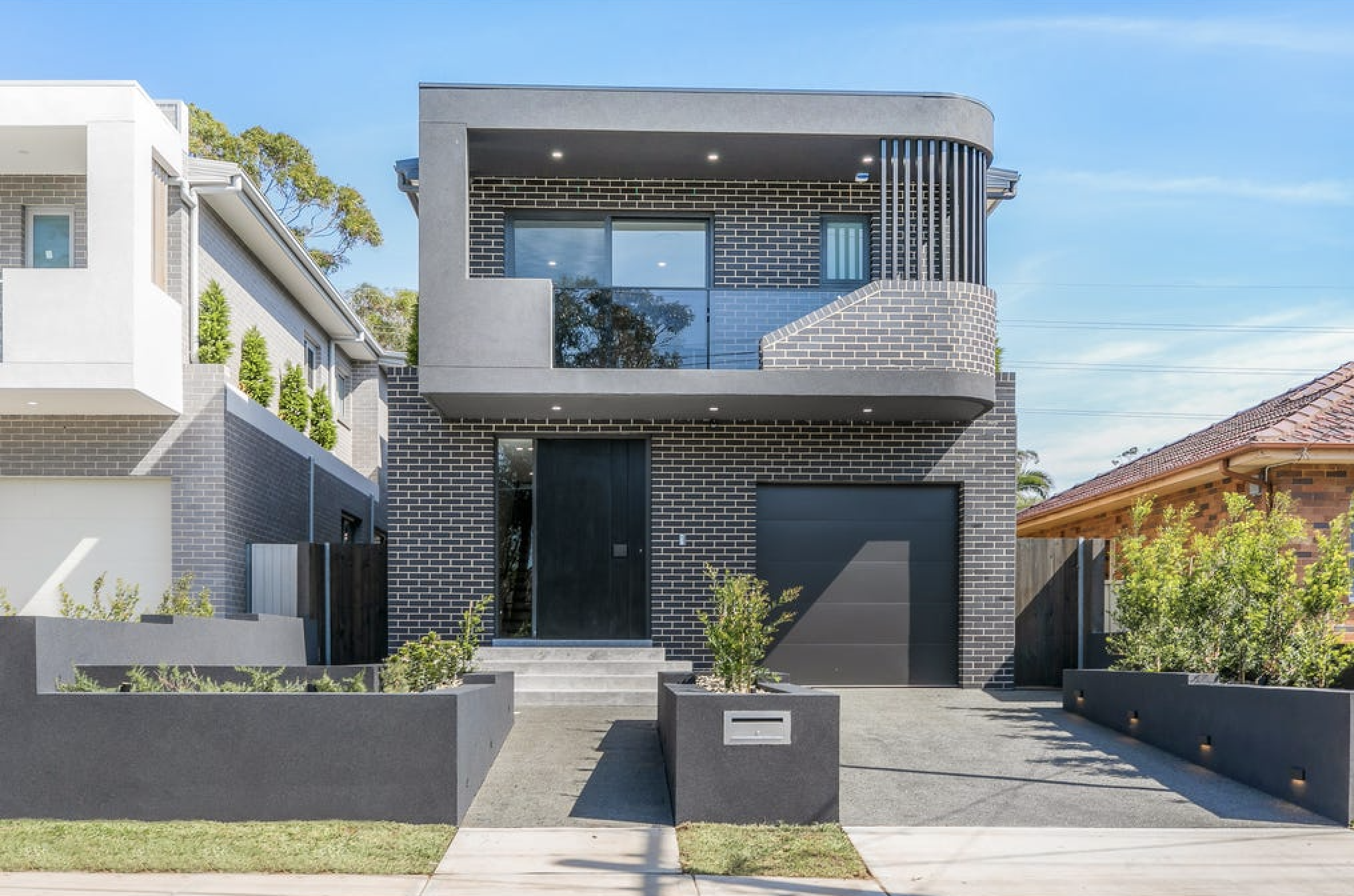 Modern two-story house with black brick facade, large windows, and a balcony, surrounded by a landscaped yard with plants and concrete planters under a clear blue sky.