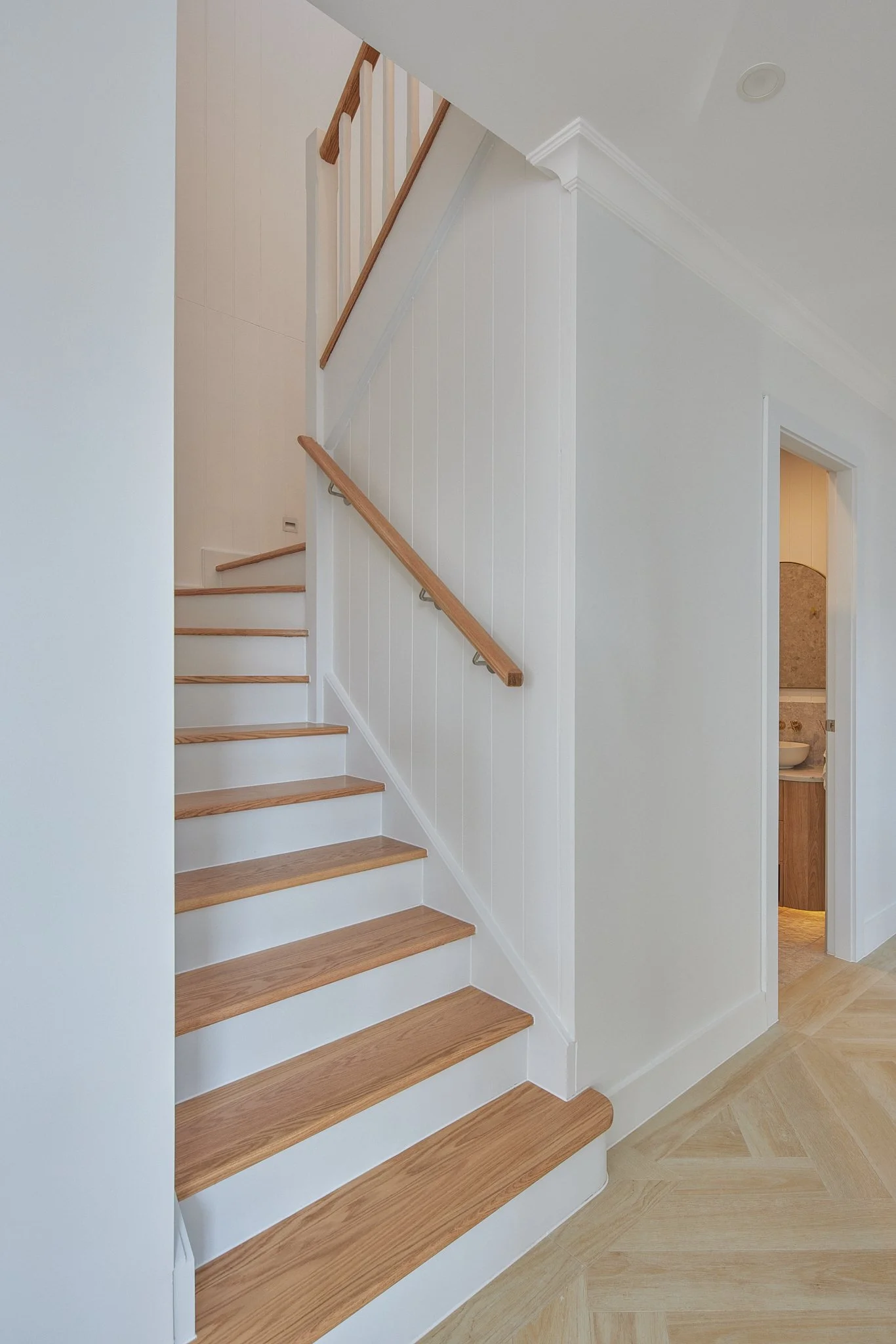 Interior view of a staircase with wooden steps, white walls, and a wooden handrail. A small bathroom with a sink is visible to the right.