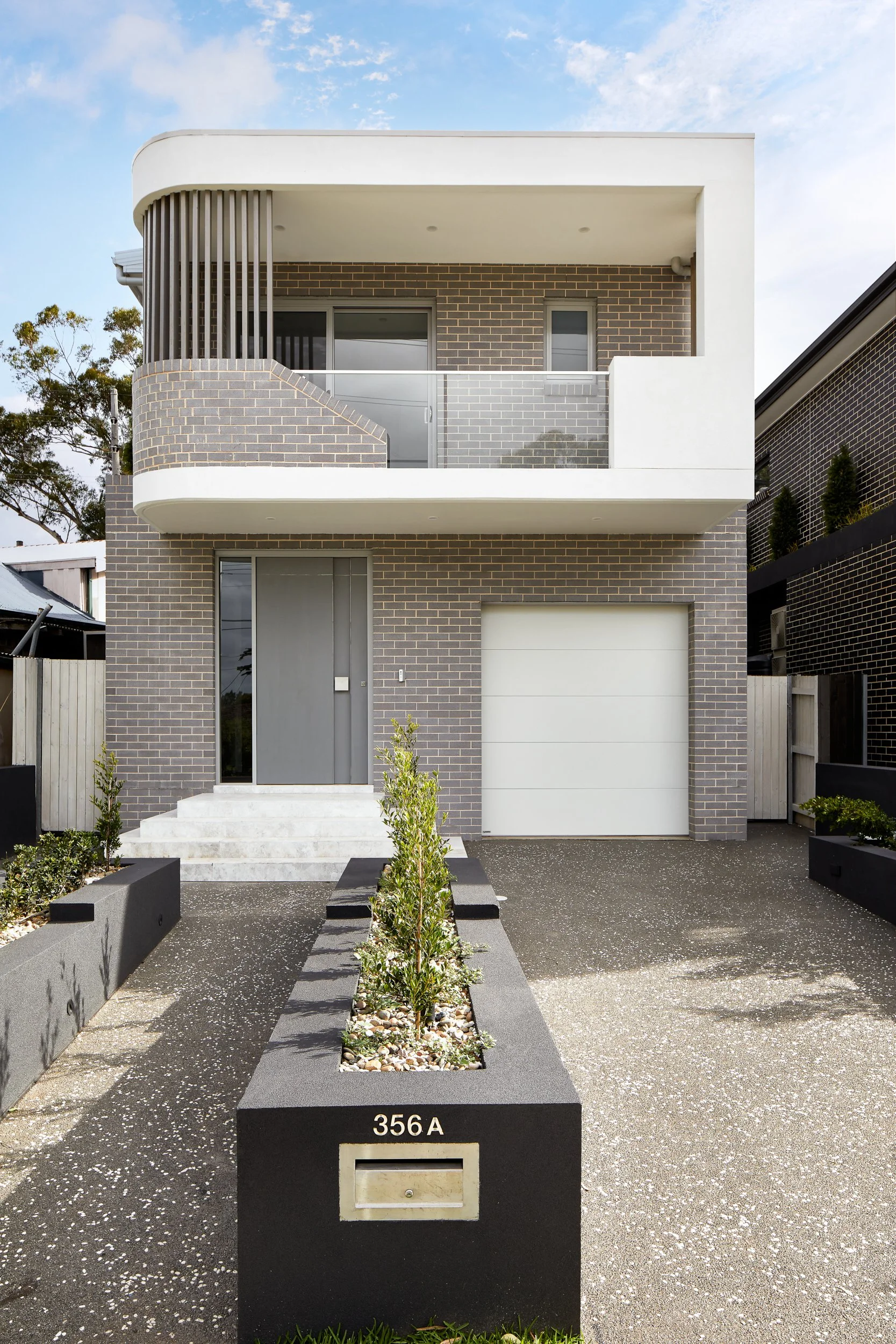 Modern two-story house with brick facade, white balcony, and gray front door, numbered 356A, with a small garden and driveway.