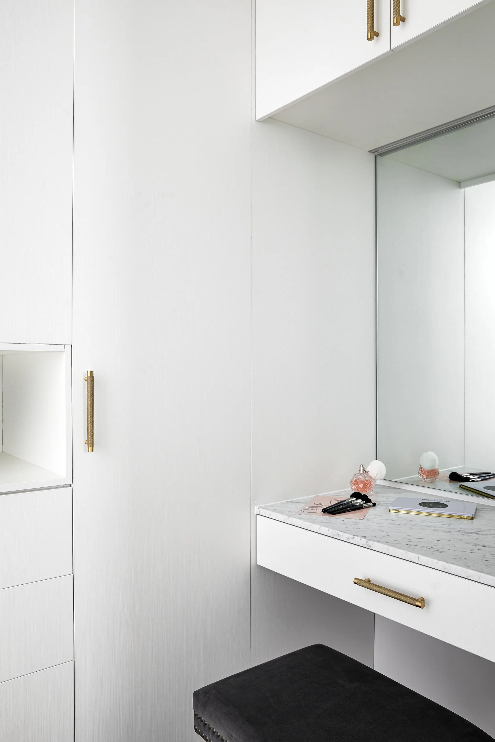 A white vanity table with makeup brushes, a notebook, and decor items in front of a large mirror, with white cabinets and a black stool.