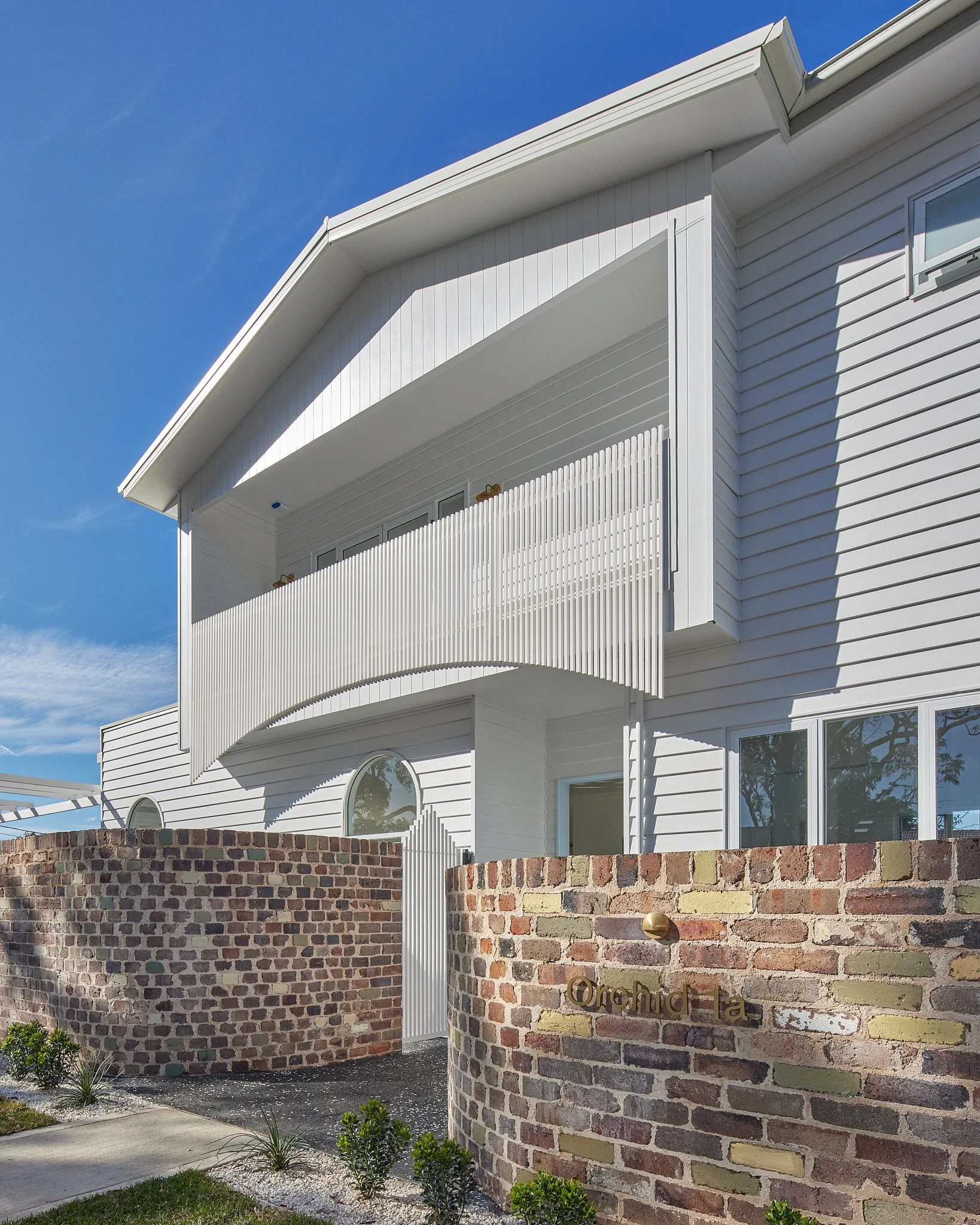 White modern house with a curved wooden balcony railing, brick wall with the house name 'Orchid Ta', small garden with plants, blue sky.