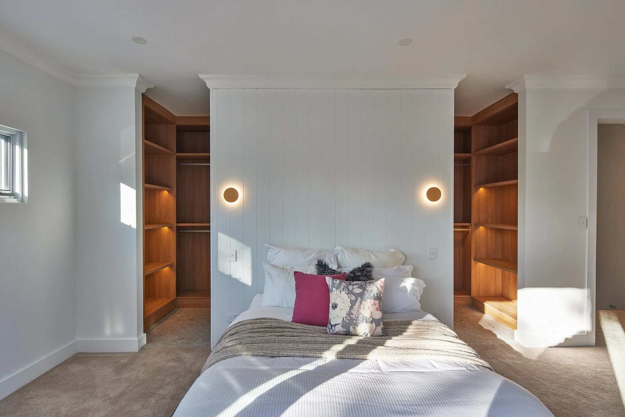 A bedroom with a bed against a white paneled wall, flanked by built-in wooden closets on each side. Two circular wall-mounted lights are on the wall, with sunlight streaming in and casting shadows on the bed. The bed has white pillows, a pink pillow,