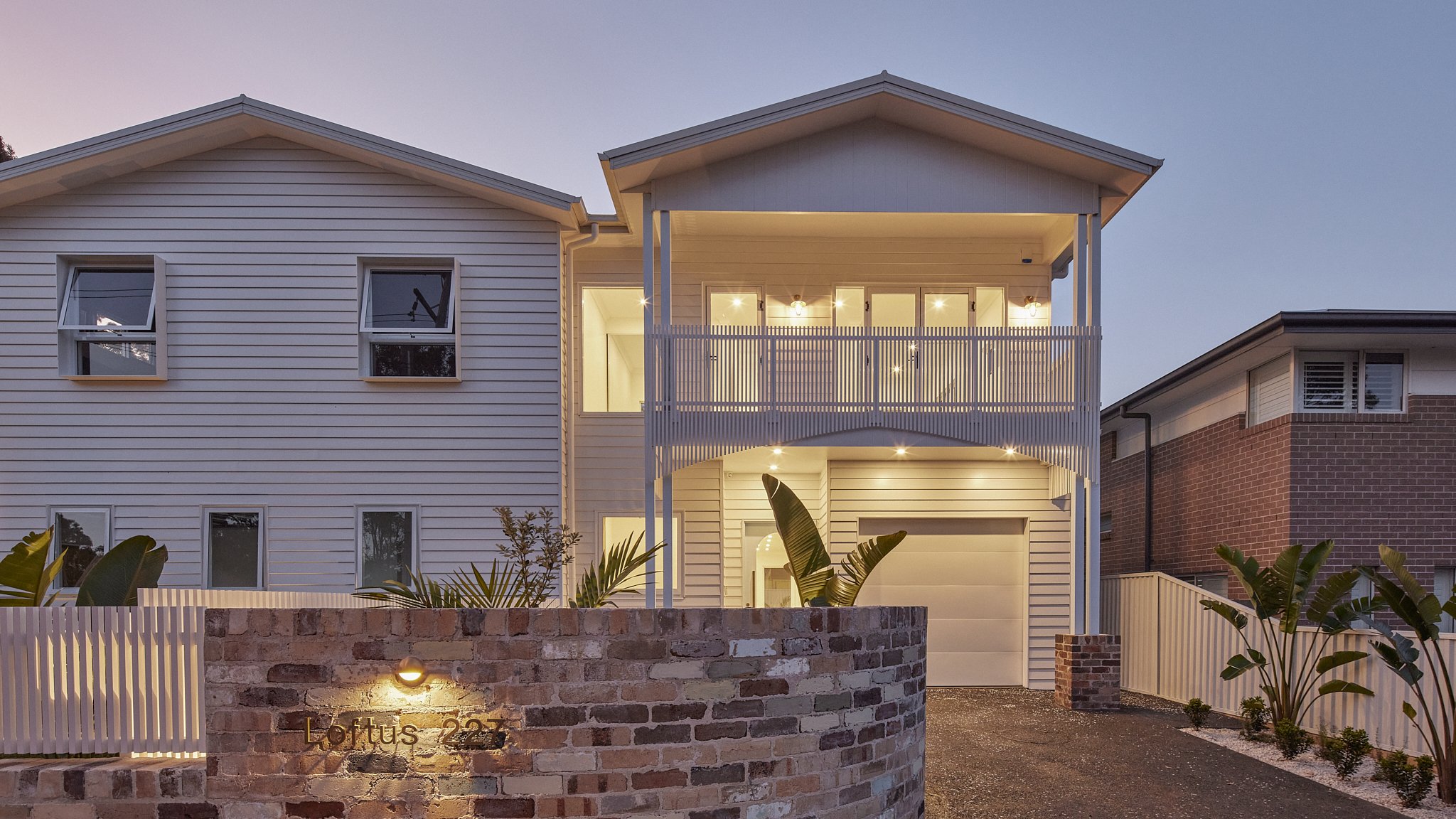 Modern two-story house with white siding, large windows, and a front garage, illuminated at dusk with outdoor lighting, brick and wood fencing, and tropical plants in the yard.