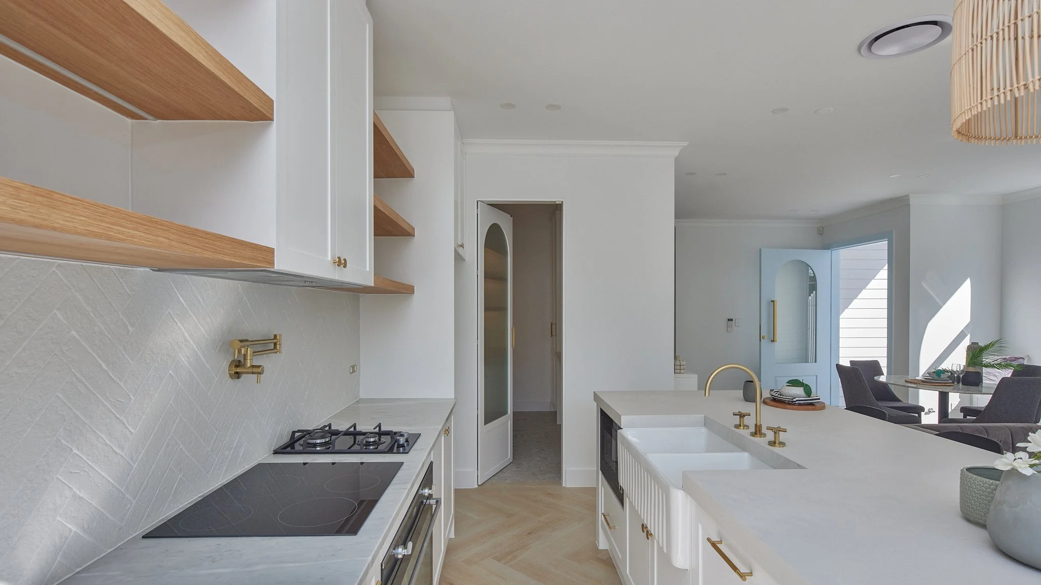 Modern kitchen with white countertops, gold fixtures, wooden open shelves, and a view into the dining area with a table and chairs near a door leading outside.