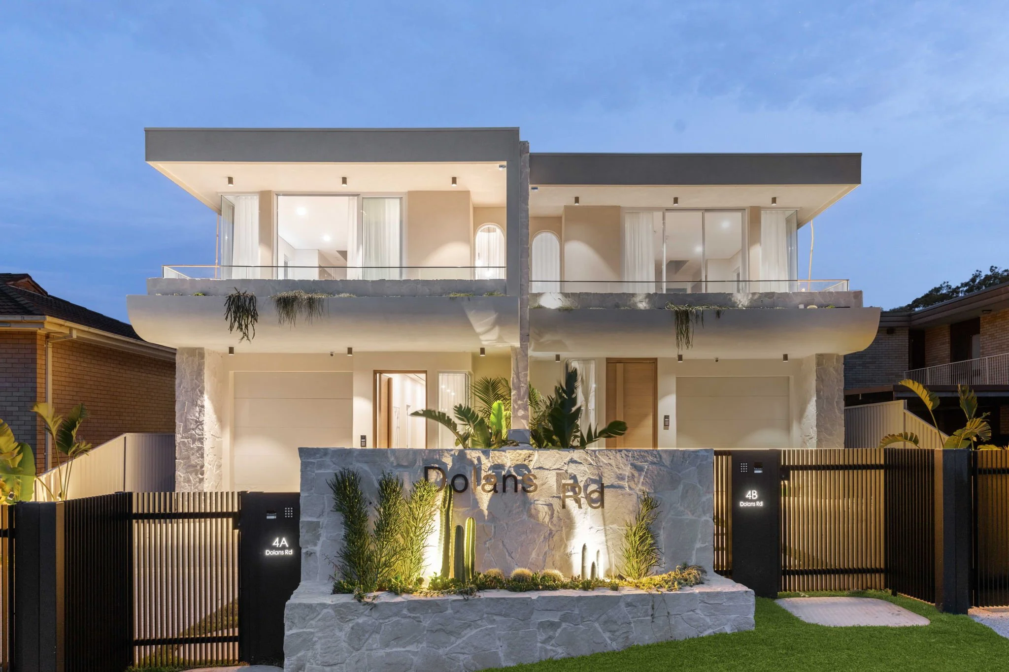 Modern multi-story house with a front yard and illuminated sign reading 'Dolans Rd', featuring a stone wall, gate, and landscaping, with a sky in the background.