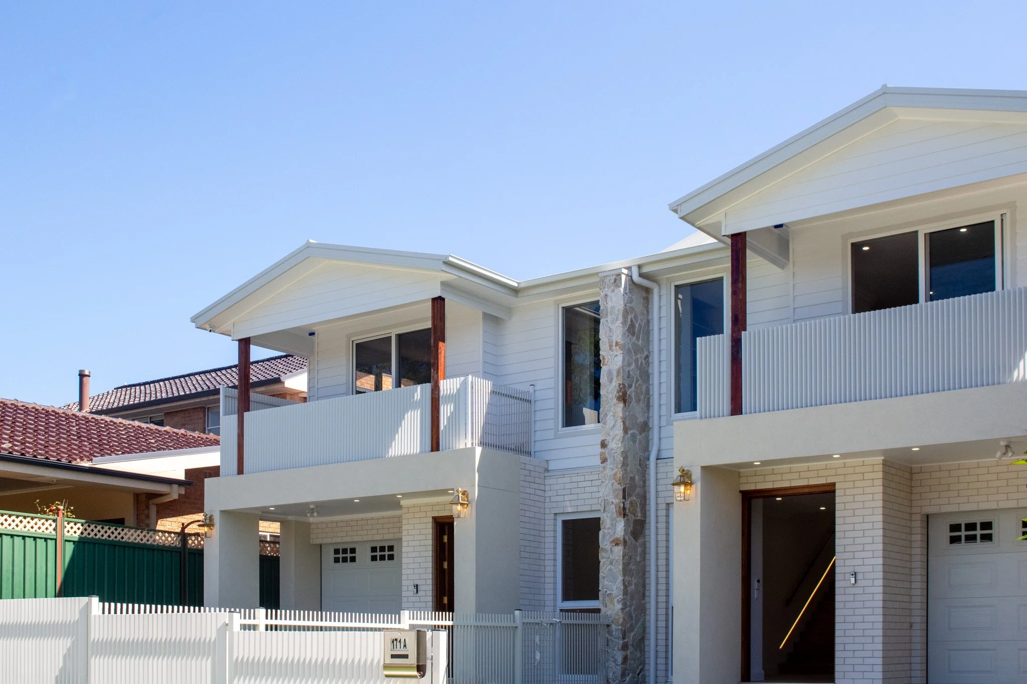 Modern residential building with white exterior, stone accent wall, two balconies with railing, and garage doors, under a clear blue sky.