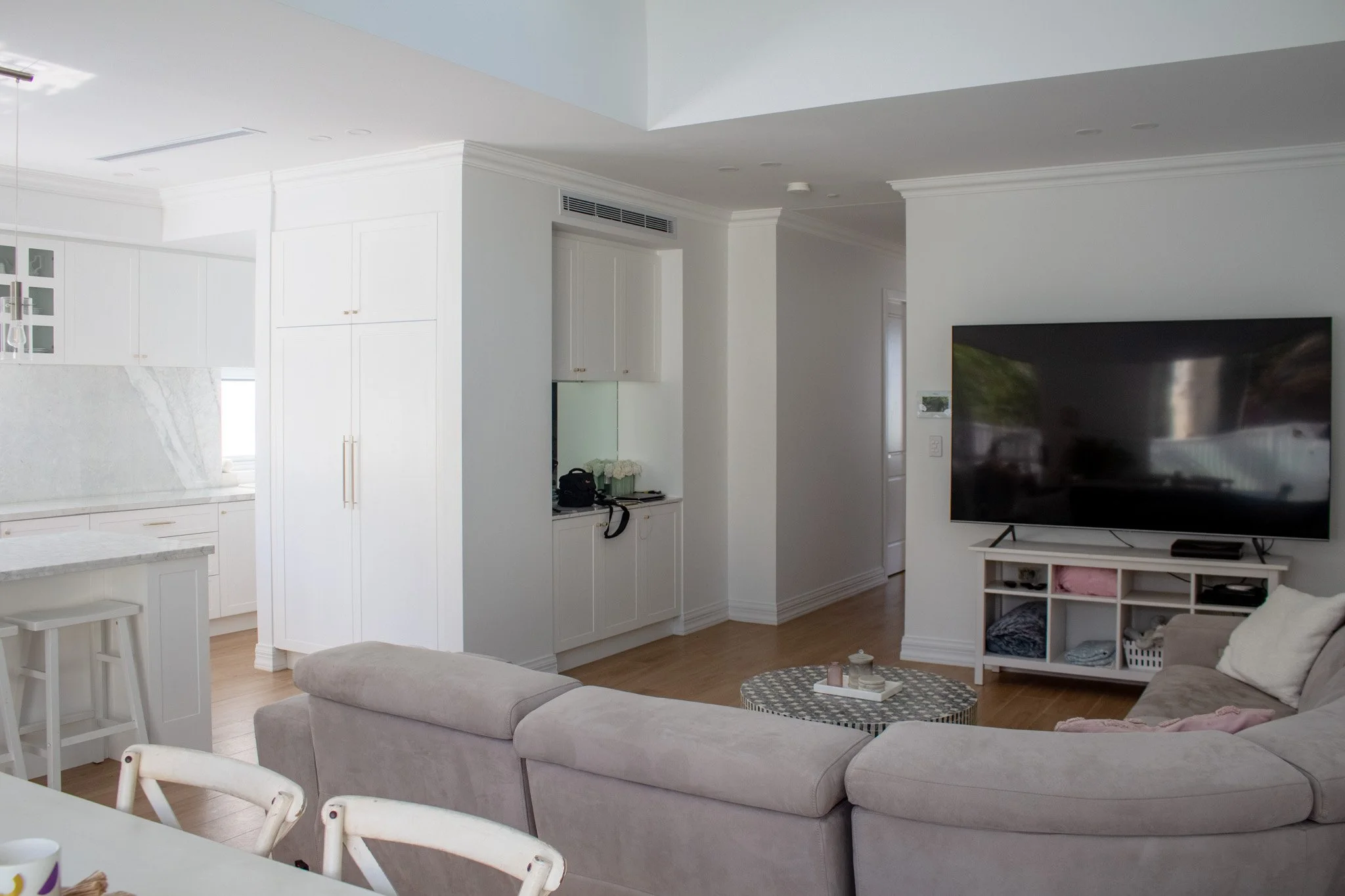 Living room with a beige sectional sofa, a small round coffee table, and a large flat-screen TV on a white stand. The background shows an open kitchen with white cabinetry and marble countertops.