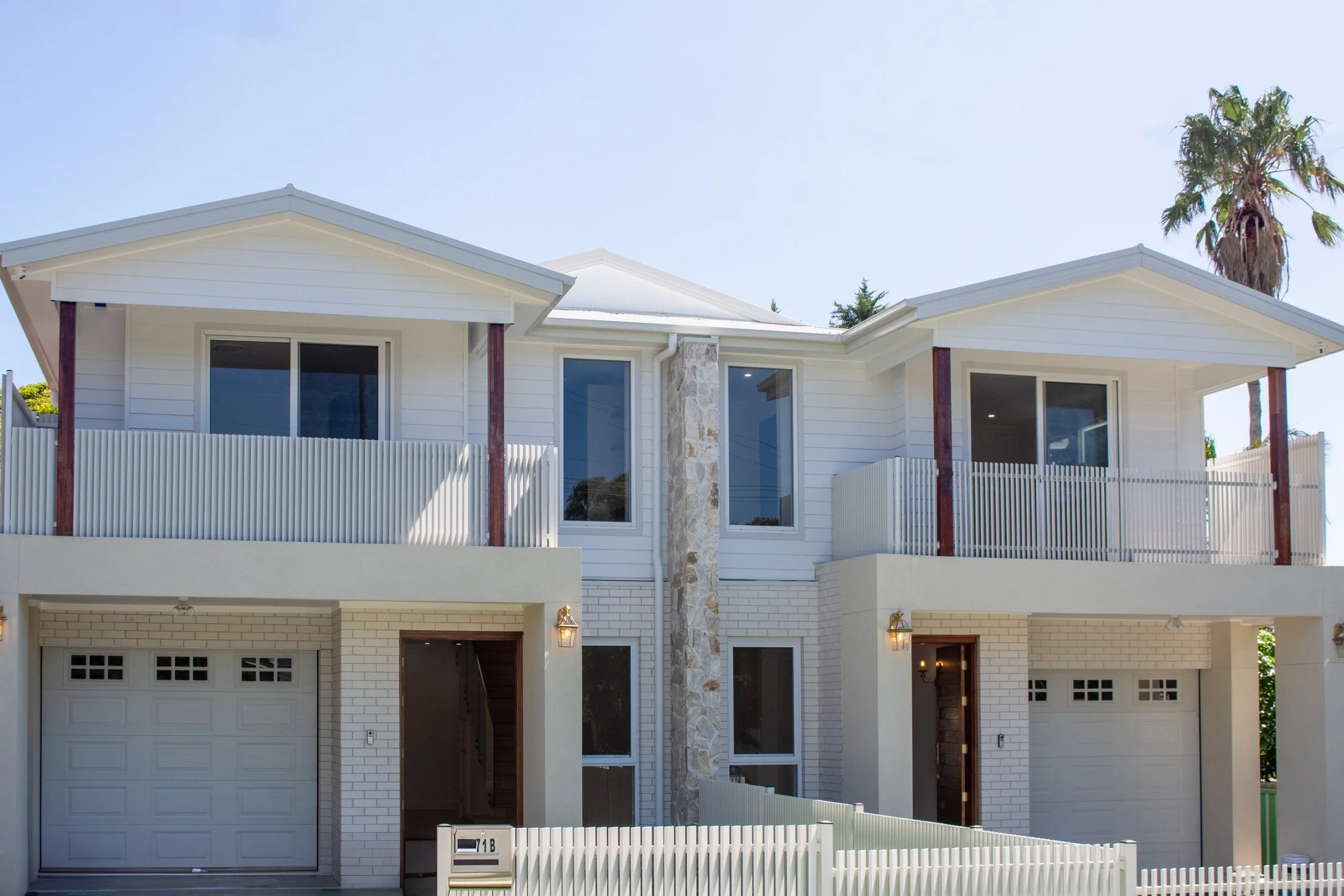 Front view of a modern two-story white house with balconies, large windows, garage doors, and a white picket fence with a palm tree in the background.