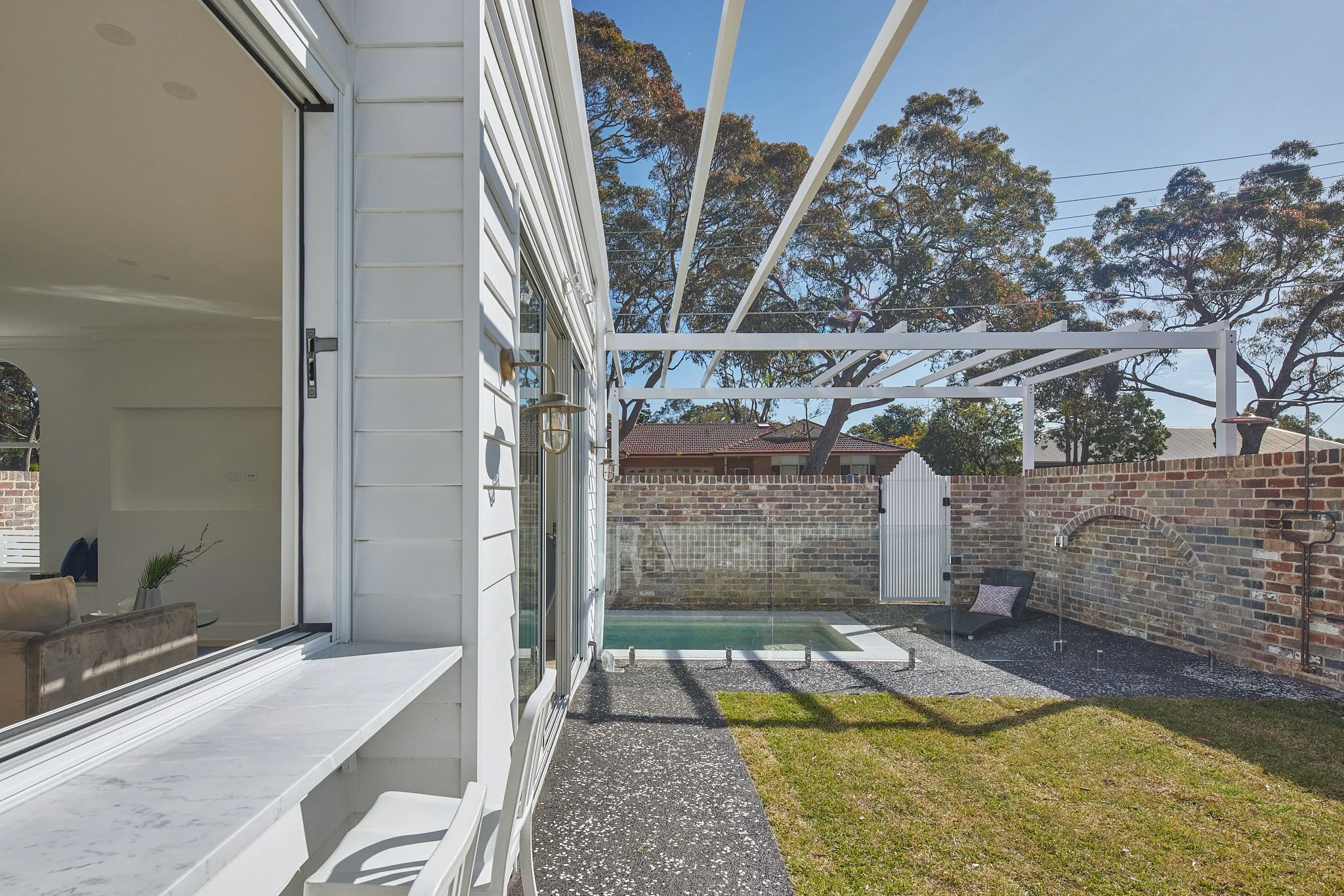 Backyard patio with a small pool, brick wall, glass fence, and outdoor shower, with trees in the background and a clear sky.