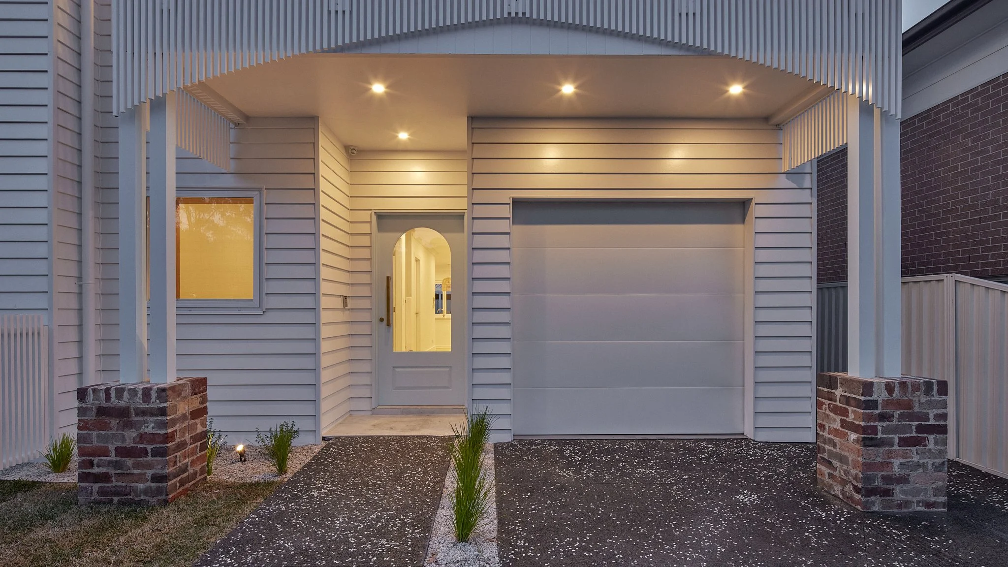 Front view of a modern house with white siding, a garage door, a white entry door with glass panel, and a small garden with plants and bricks.