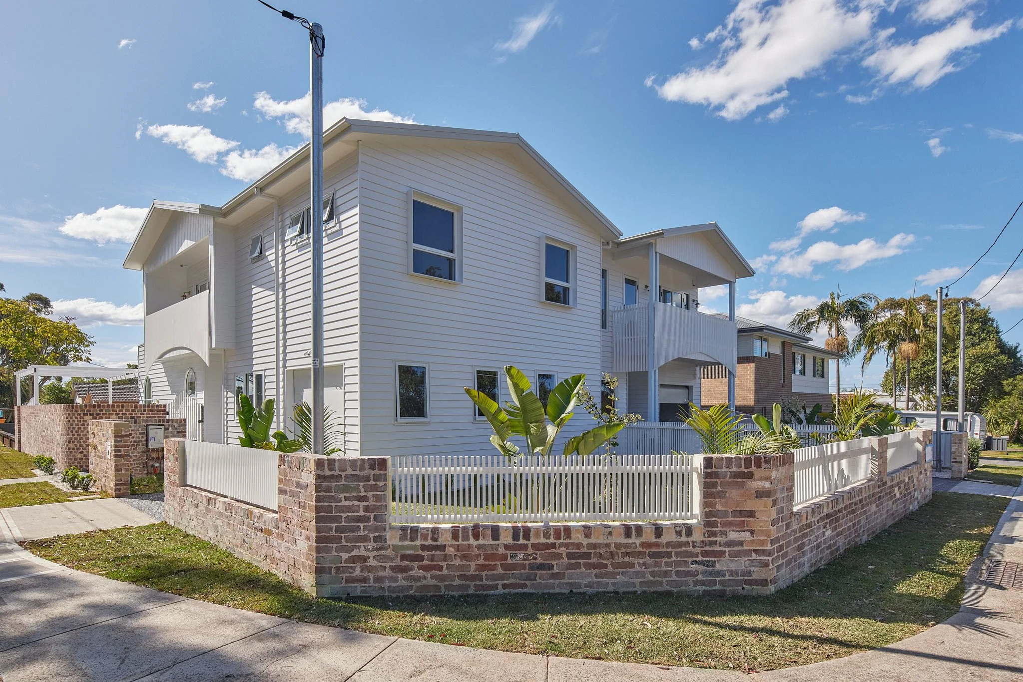 A white multi-story house with a brick fence, tropical plants, and a sidewalk, under a partly cloudy sky.