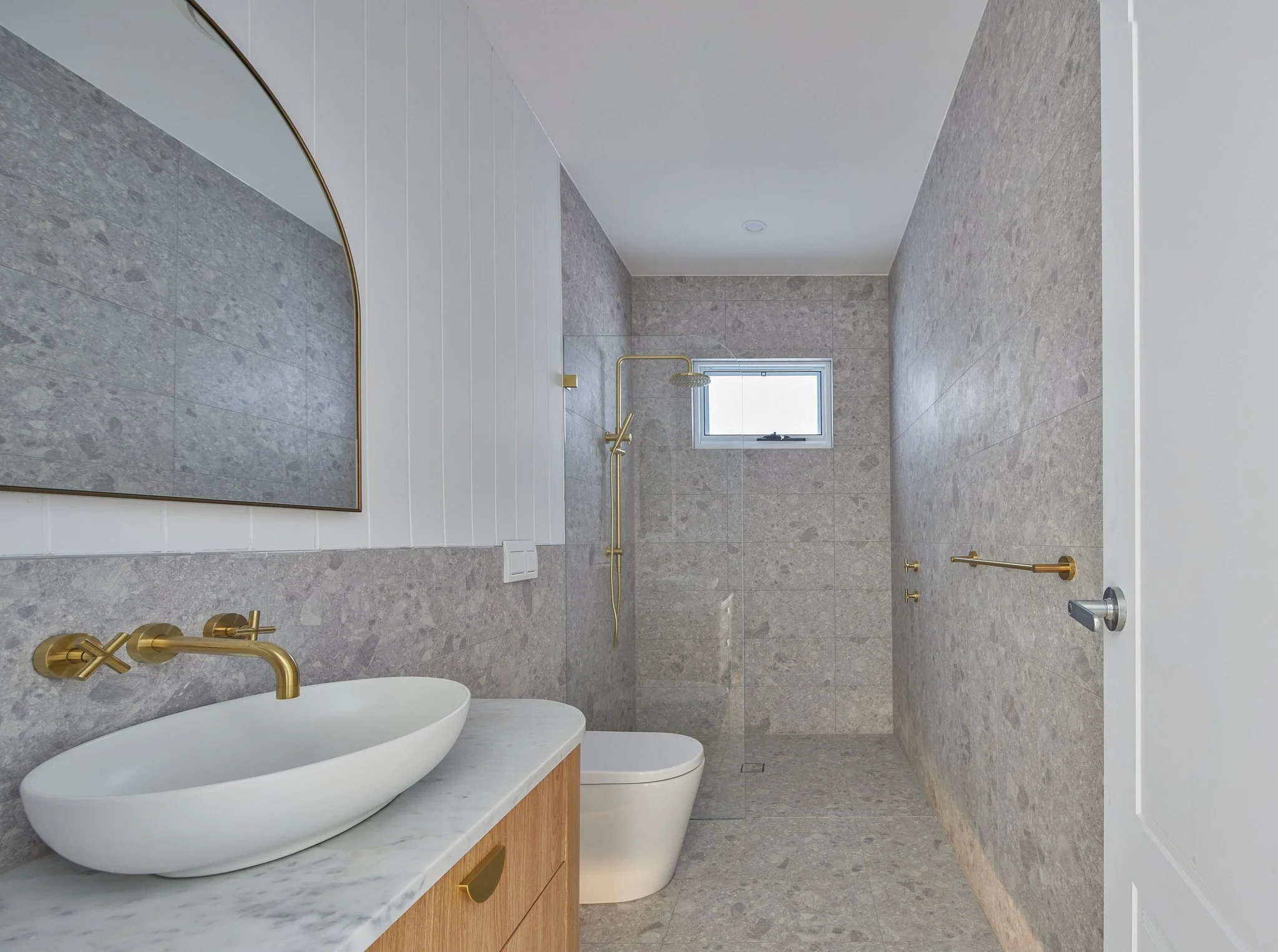 Modern bathroom with gray stone walls and floor, a white door, a wooden vanity with a white vessel sink and gold faucet, a toilet, a small window, a gold showerhead and horizontal bar, and a large mirror.