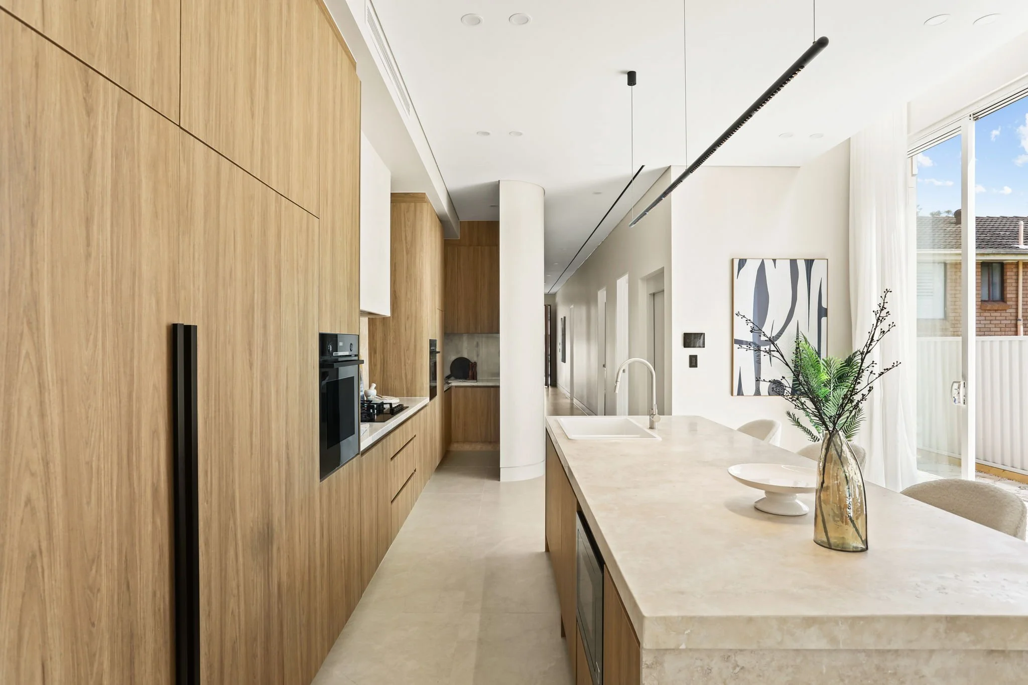 Modern kitchen with wooden cabinets, light-colored island, and a glass door leading to a balcony with blue sky and neighboring building visible.