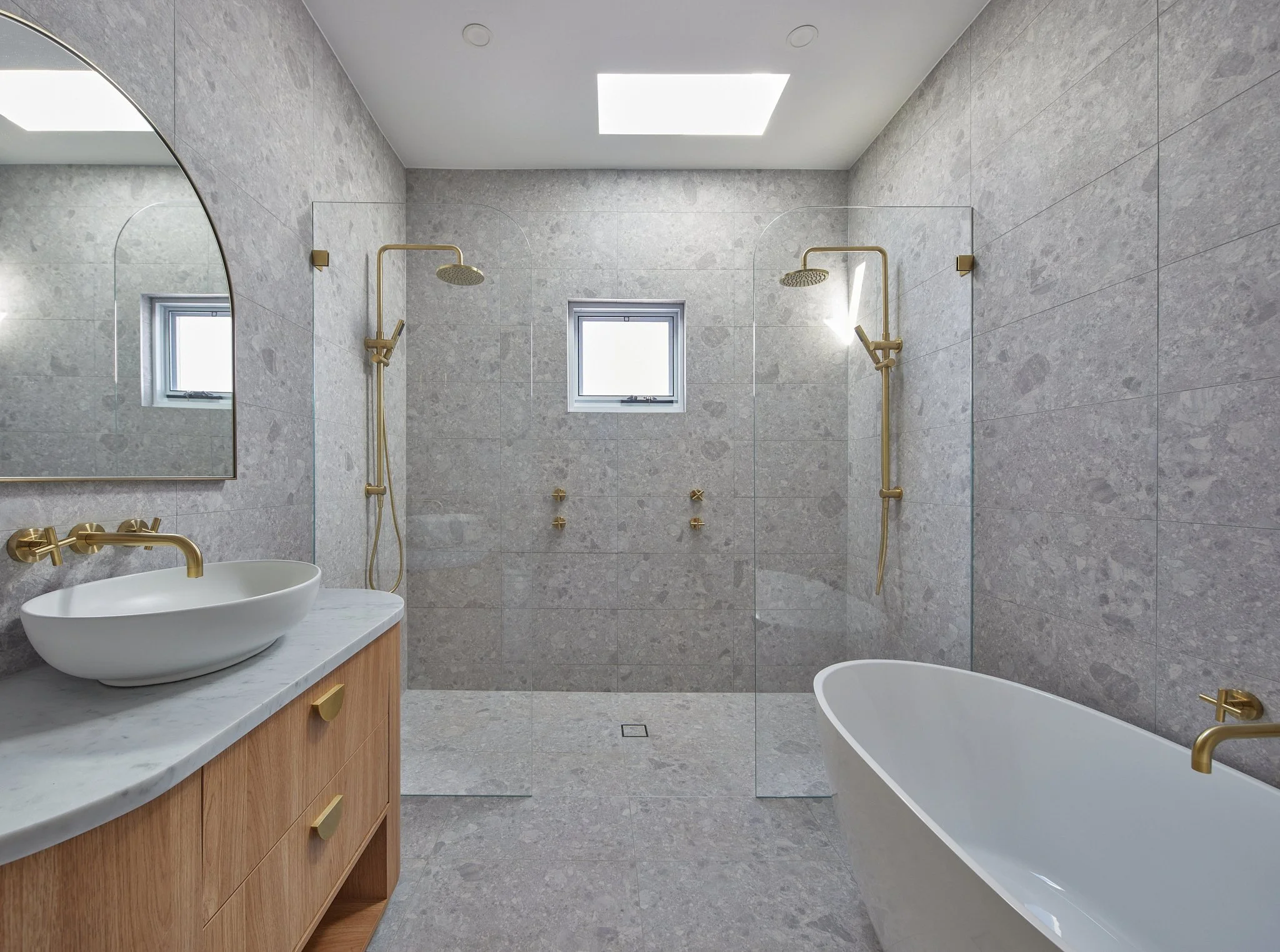 Modern bathroom with gray stone walls, a freestanding bathtub, a wooden vanity with a vessel sink, gold fixtures, two showerheads, and two small windows.