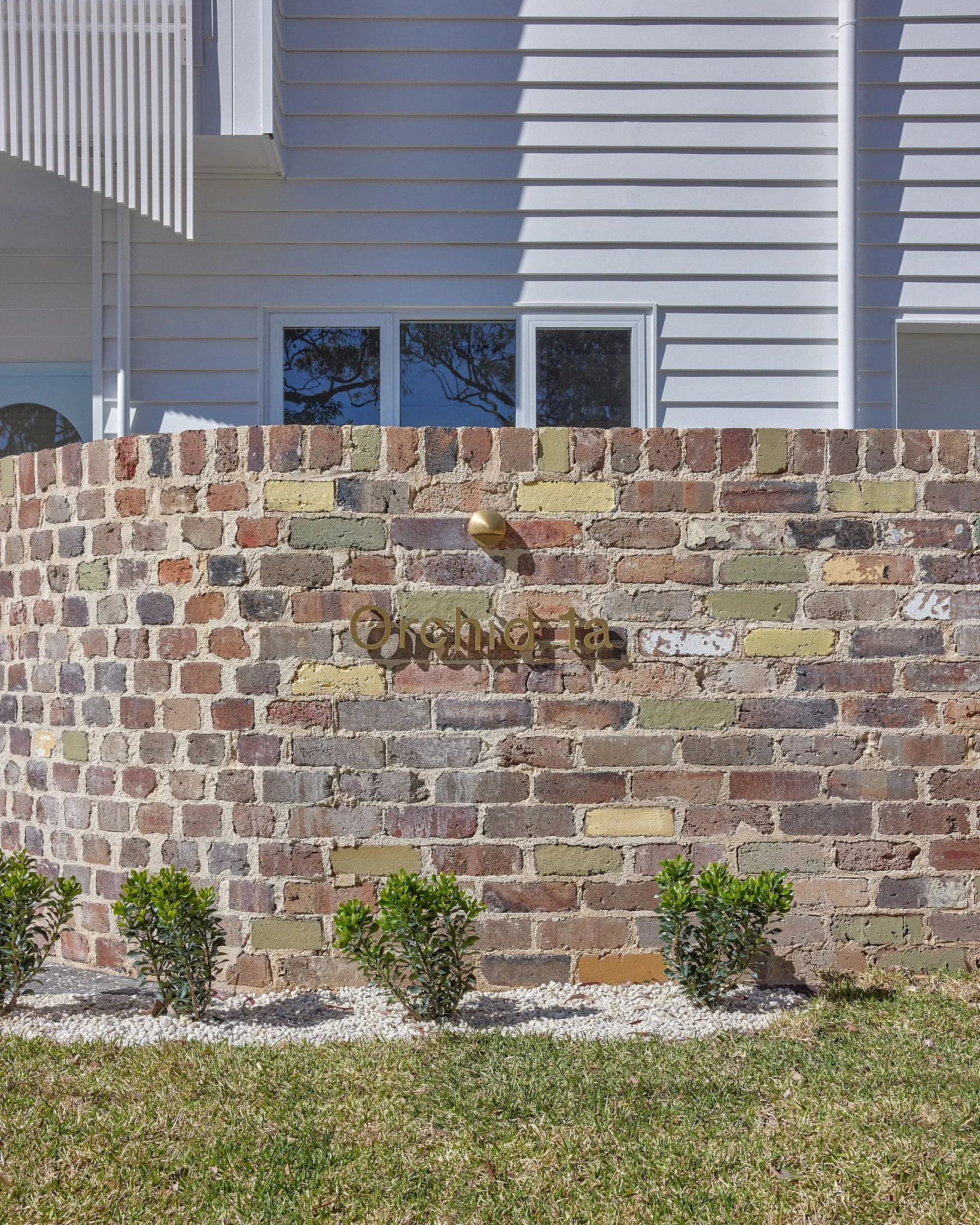 A brick wall with the word "Orchida" on it and a small sculpture above it. There are small bushes at the base of the wall, and in the background, there is a white house with windows.