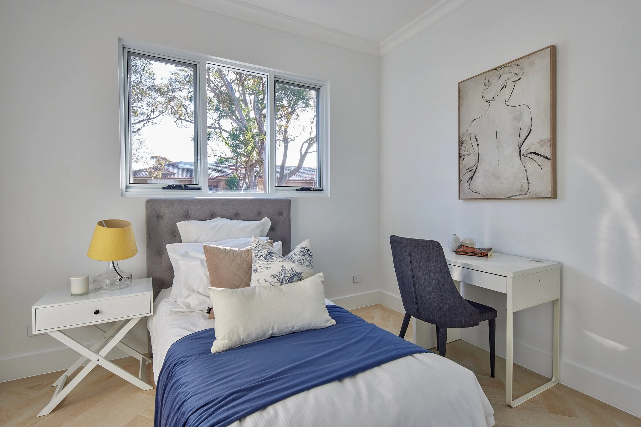 A bedroom featuring a bed with white and beige pillows, a blue blanket, and a grey upholstered headboard. There's a white nightstand with a yellow lamp and a candle. A window behind the bed shows trees outside. To the right, there's a white desk with