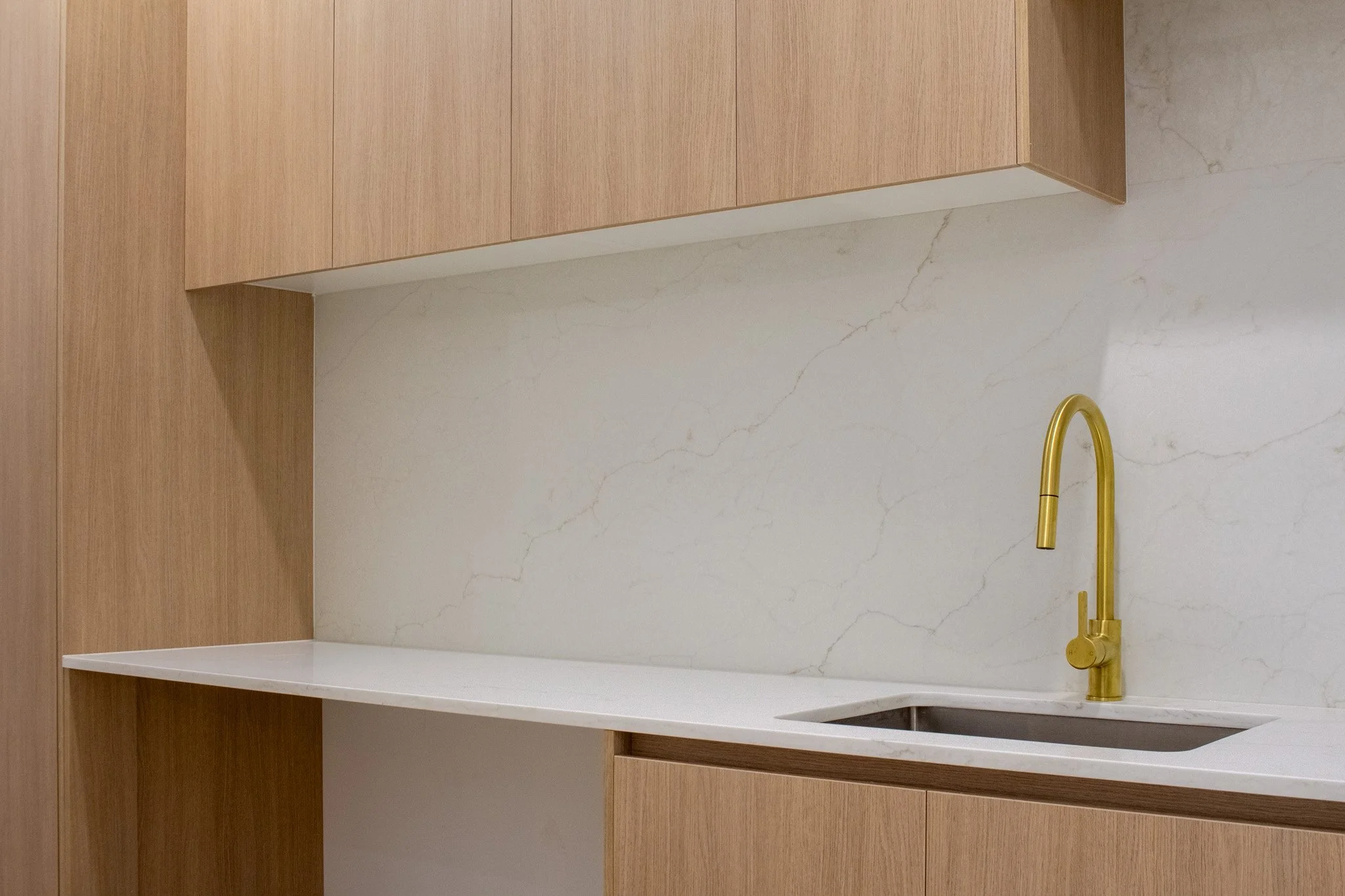 Modern kitchen with a white marble backsplash and countertop, wooden cabinets, and a gold faucet over a small stainless steel sink.
