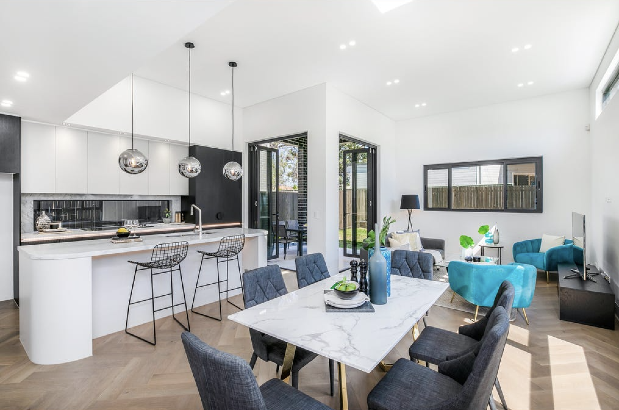 Open-concept living room and kitchen with white walls, modern furniture, a marble dining table, blue and grey chairs, and large windows letting in natural light.