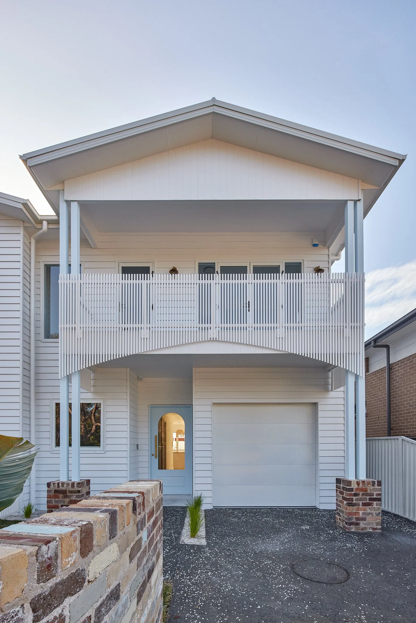Modern two-story white house with a garage, balcony with vertical slat railing, and a curved brick wall at the front.